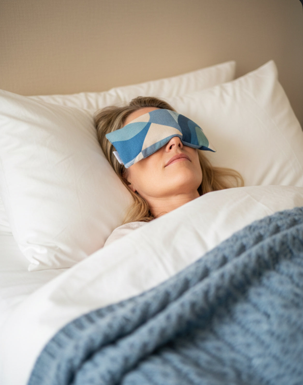 A women laid in bed with a multi blue abstract eye pillow across her eyes resting under white cotton sheets and a blue woollen throw