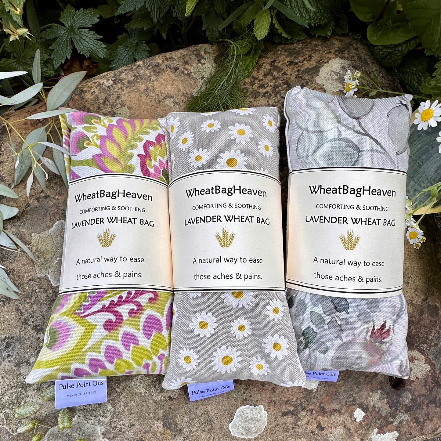 Three in a row lovely cotton wheat bags with dried lavender buds for holistic relaxation. From left to right Nikita, bright and cheery greens and pink floral, then a lovely Daisy print wheat bag and lastly a watercolour botanical print in muted greys and pinks, pictured in the garden at WheatBagHeaven backdrop 
