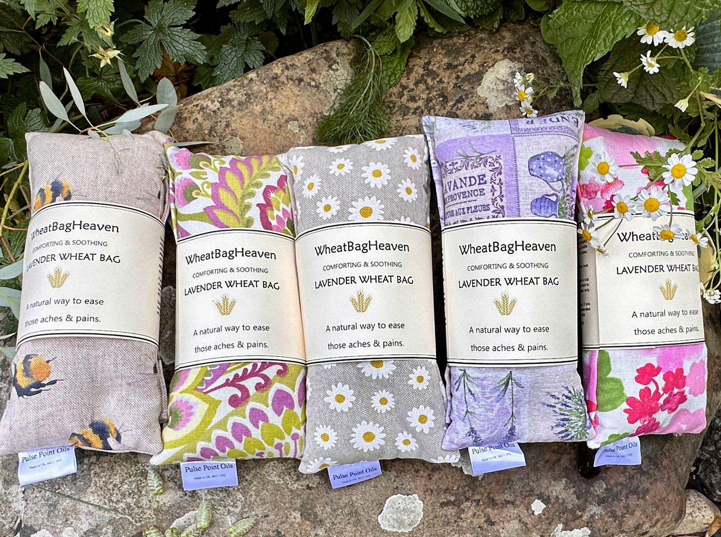 Five in a row, lavender scented wheat bags from WheatBagHeaven.com A garden backdrop from left to right, yellow bumblebee print wheat bag then Nikita, bright mood lifting floral print heat wrap, Daisy flower printed wheat bag, followed by lavender patchwork, a cotton print depicting all that’s French lavender, floral bouquets and chateaux and lastly beautiful pink peony printed wheat bag. Filled with Somerset whole wheat and English lavender buds for holistic wellbeing 