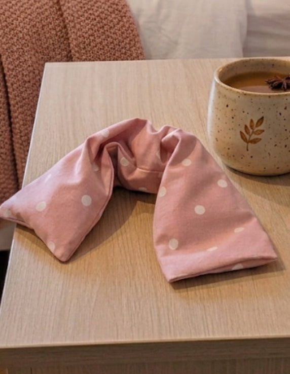 Close up of Pink with off white dots lavender wheat bag from WheatBagHeaven. The wheat bag is placed on a bedside table next to a mug of coffee and journaling book and pen. Bed draped in pink knitted throw and cushions