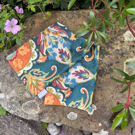 psychedelic floral cotton orinted cover for wheat bag in orange, teal, blue and green colour way. pictured on a stone backdrop in the garden of wheat bag heaven