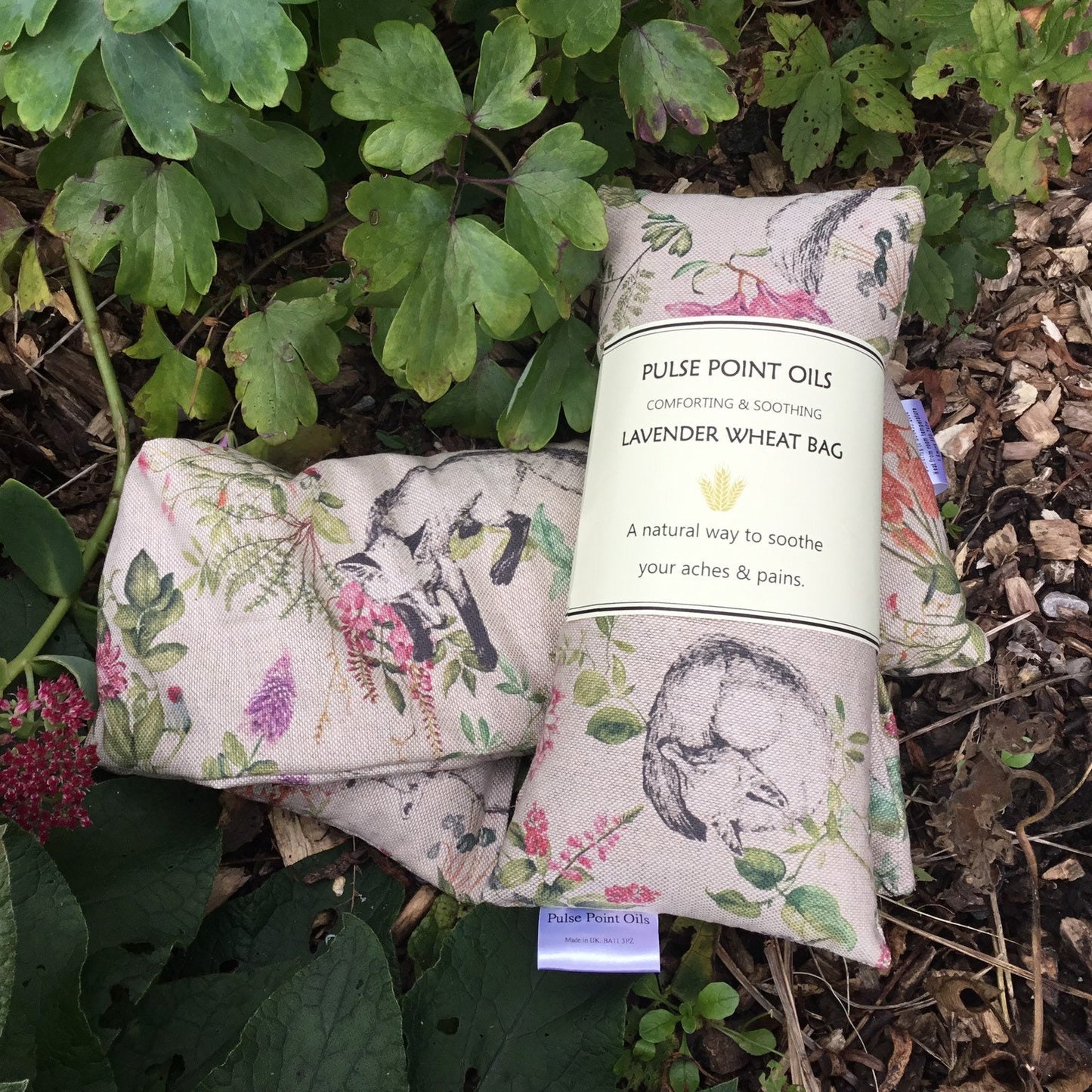 Two lavender wheat bags in an Urban fox print. The foxes are grey outlined and frolicking amongst a floral botanicals, in shades of pink and greens on a beige base