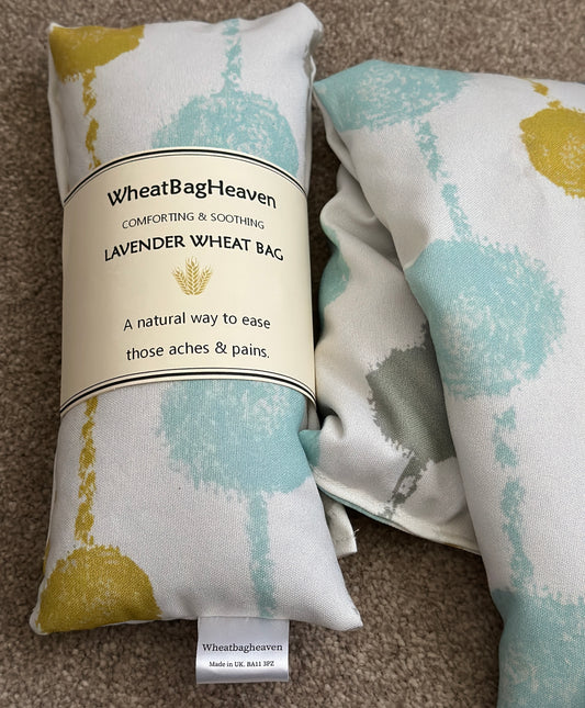 two lavender wheat bags with a Pom Pom abstract print in turquoise, blues and ochre on a light background.  One has makers belly band that reads WheatBagHeaven comforting and soothing lavender wheat bag, a natural way to ease those aches and pains 