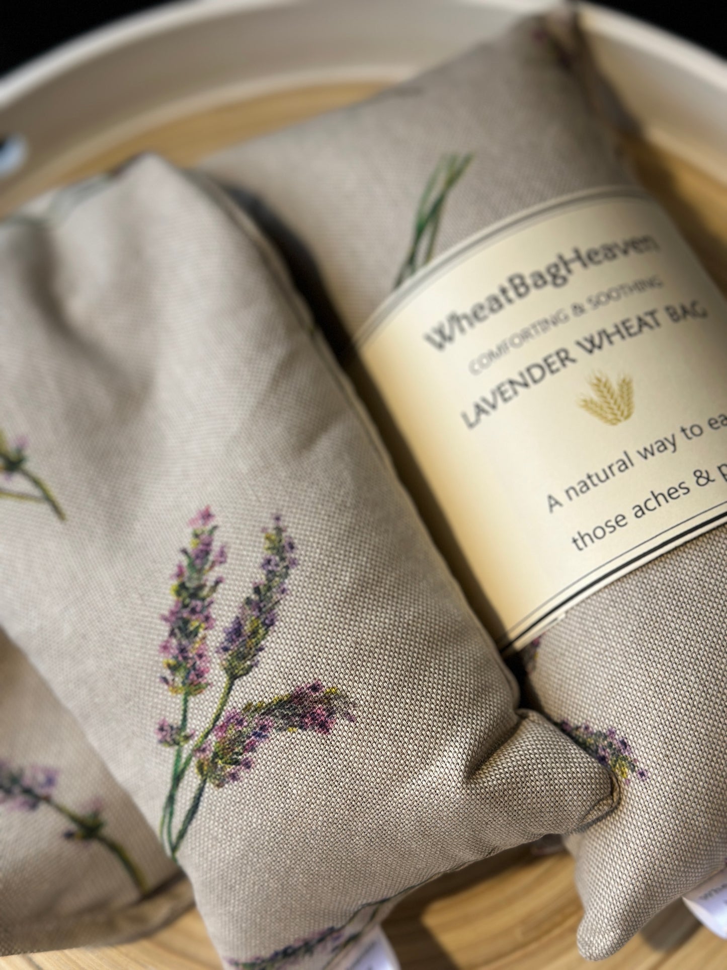 Two Wheat bags with a lavender flower print design on a wooden surface. One wheat bag has maker's belly band that read WheatBagHeaven comforting and soothing Lavender Wheat Bag A natural way to ease those aches and pains. Both have makers tags