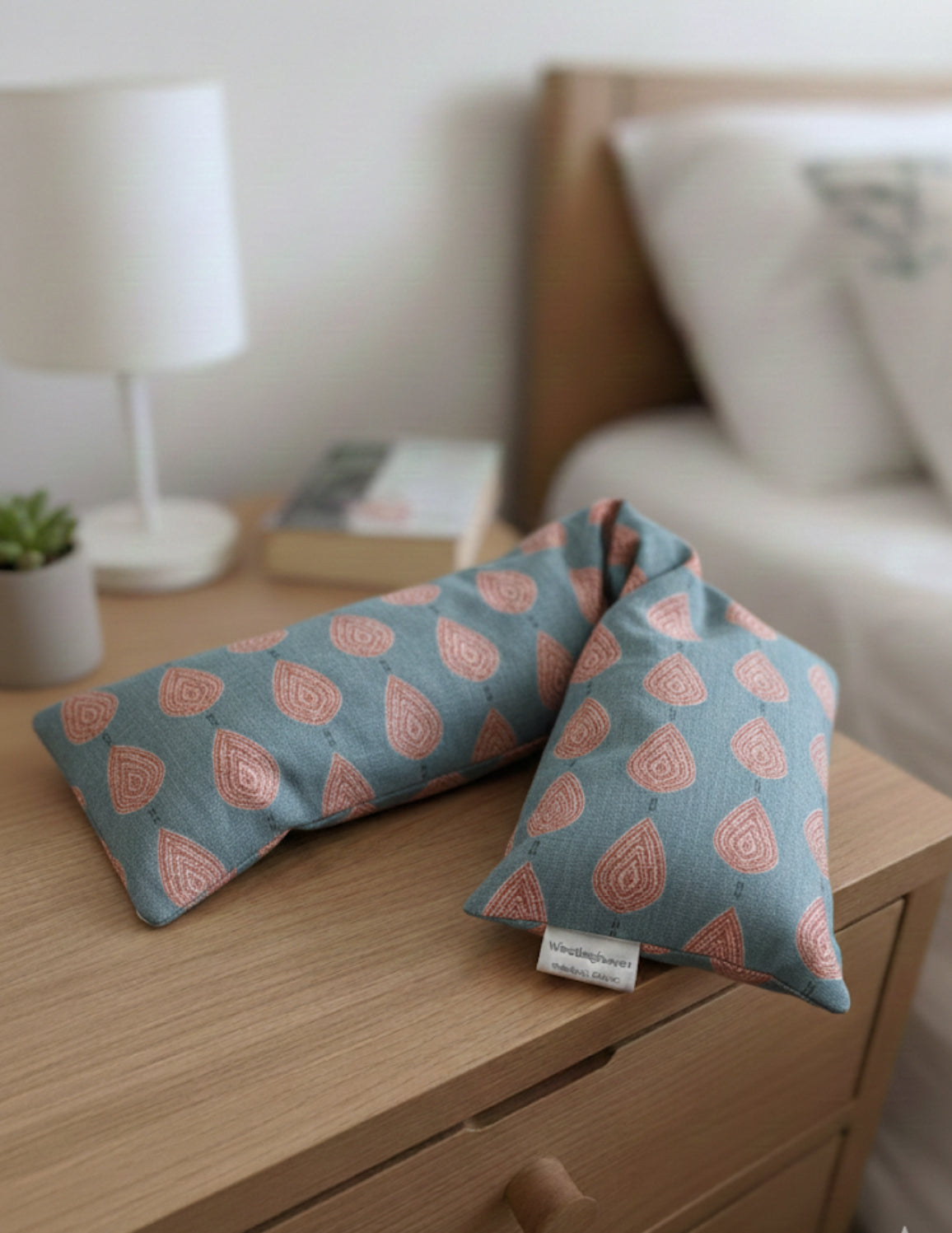 Two patterned wheat bags on a wooden surface with a blurred background of a bed. Wheat bags are in a pink paisley teardrop pattern on a soft teal base, with a  Makers tag that reads 'WheatBagHeaven'