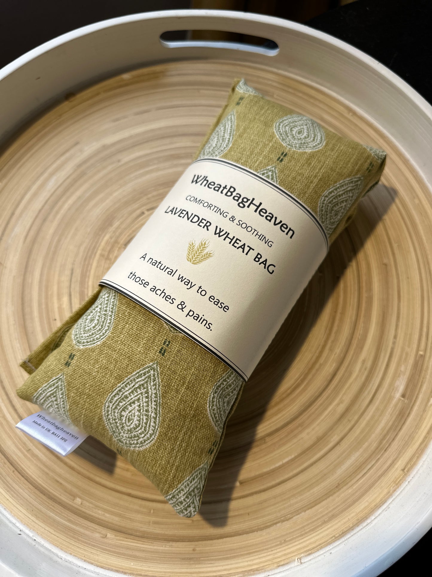 A lavender wheat bag on a wooden tray. The wheat bag has a paisley teardrop pattern in shades of green and cream with a base colour of ochre. Its labelled "WheatBagHeaven" comforting and soothing lavender wheat bag. A natural way to ease those aches and pains. Also a tag that reads WheatBagHeaven