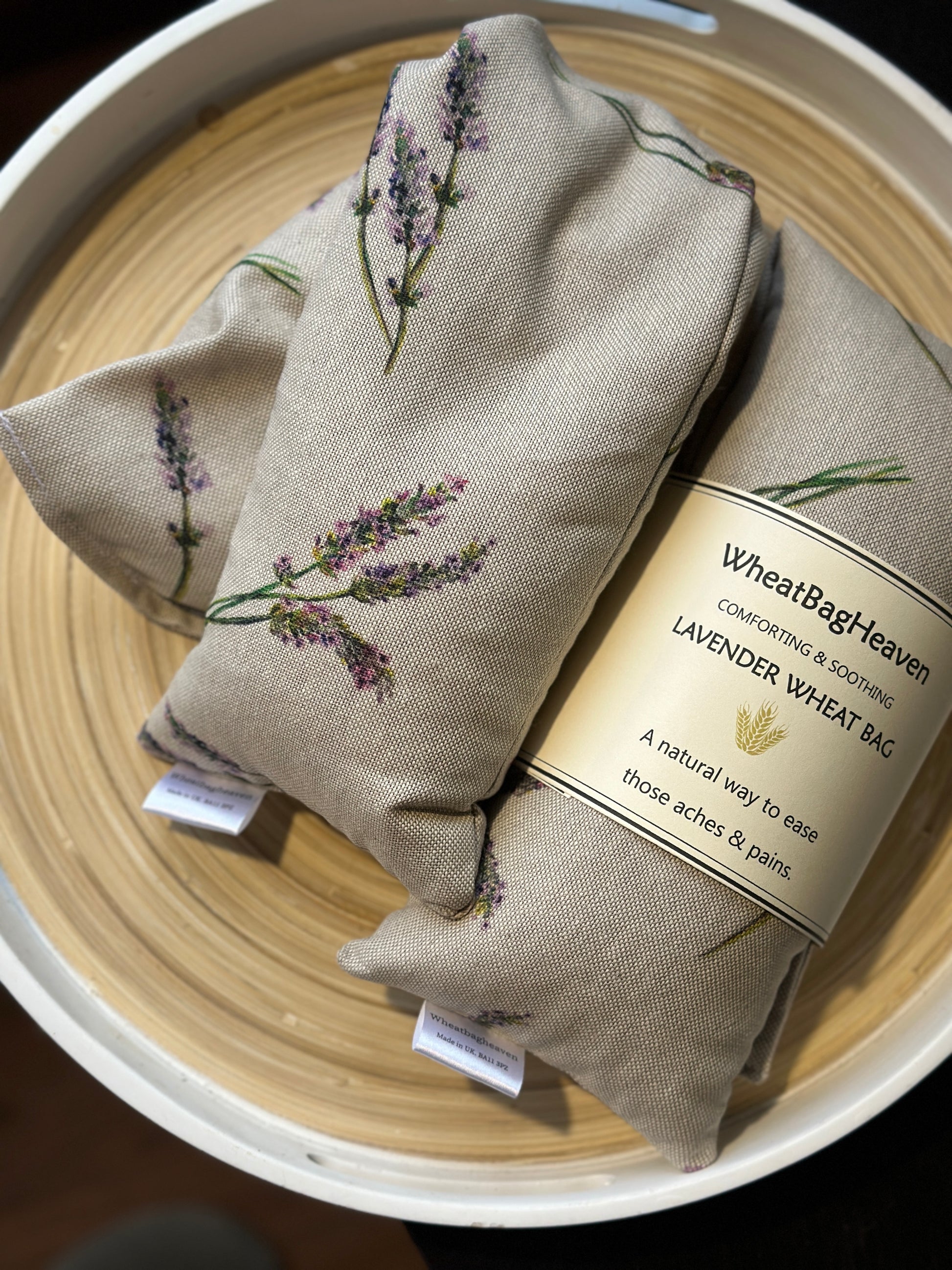 Two Wheat bags with a lavender flower print design on a wooden surface. One wheat bag has maker's belly band that read WheatBagHeaven comforting and soothing Lavender Wheat Bag A natural way to ease those aches and pains. Both have makers tags