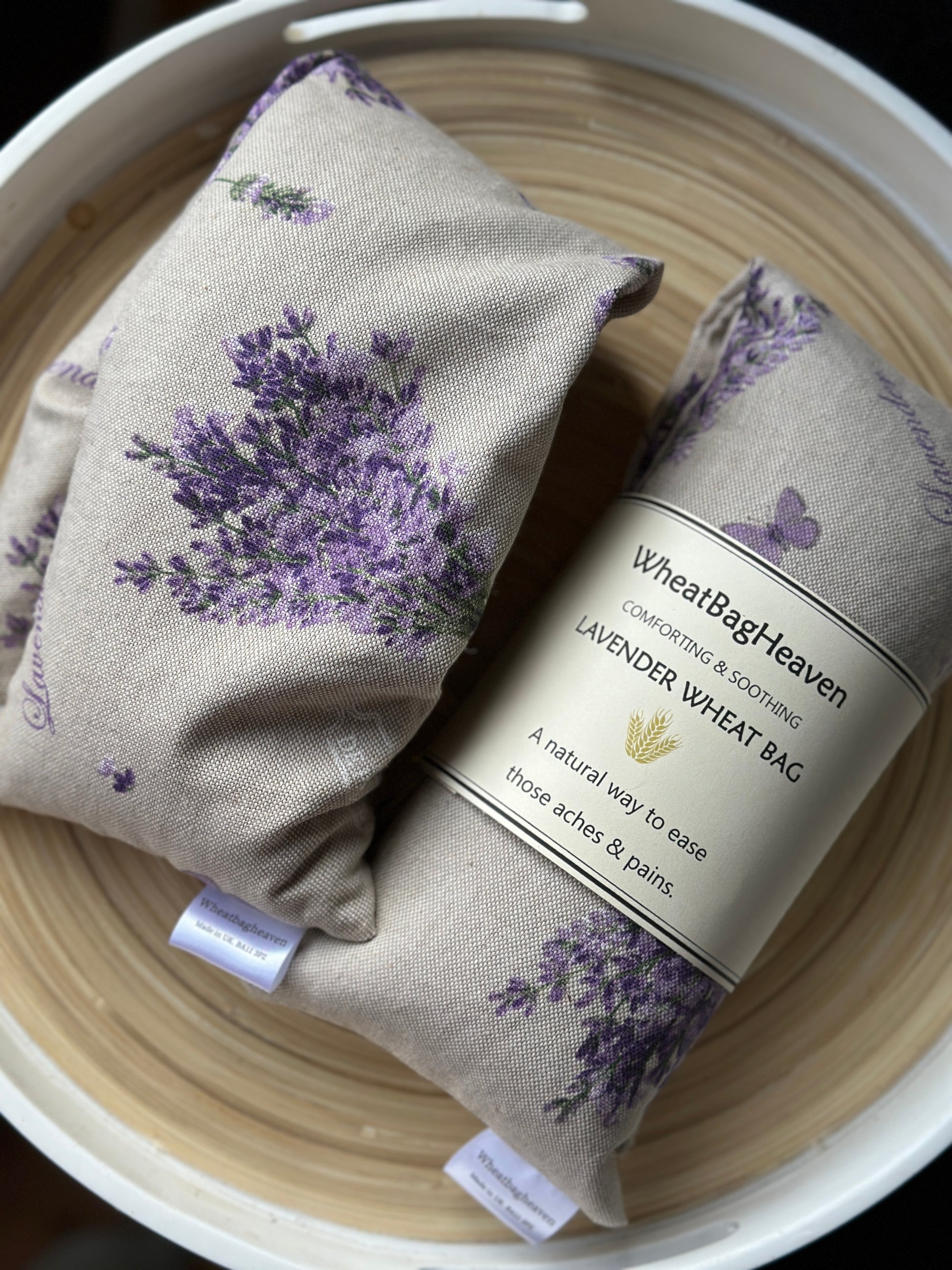 Two Wheat bag with lavender floral bouquet print and label that reads WheatBagHeaven comforting and soothing Lavender Wheat Bag. A natural way to ease those aches and pains. The wheat bags are laid in a wooden tray