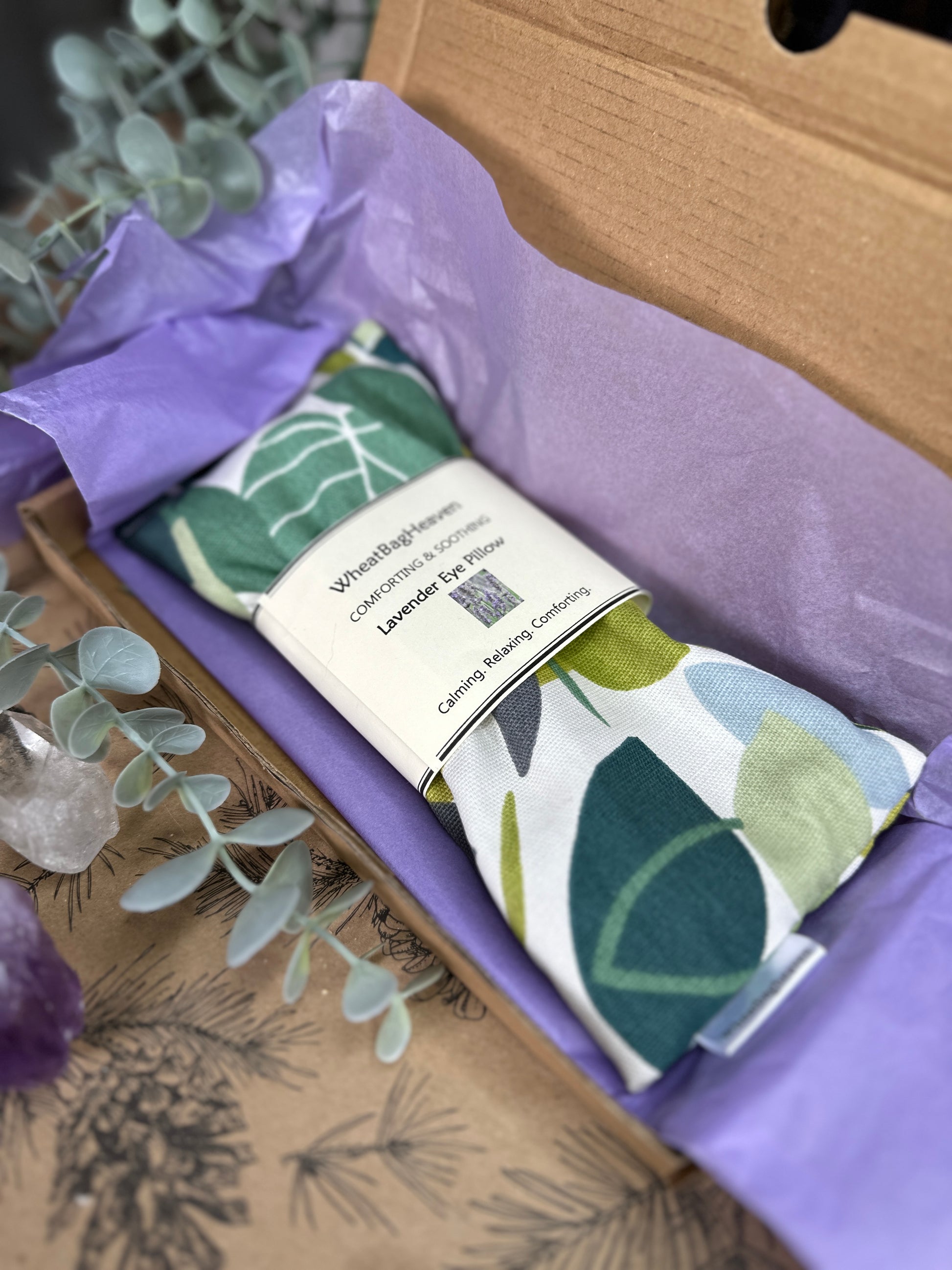 Lavender eye pillow green spruce with all over leaf pattern in various shades of green on a white base. The eye pillow is laid on purple tissue paper in a letterbox sized box. A label reads WheatBagHeaven comforting and soothing Lavender Eye Pillow, calming. relaxing, comforting