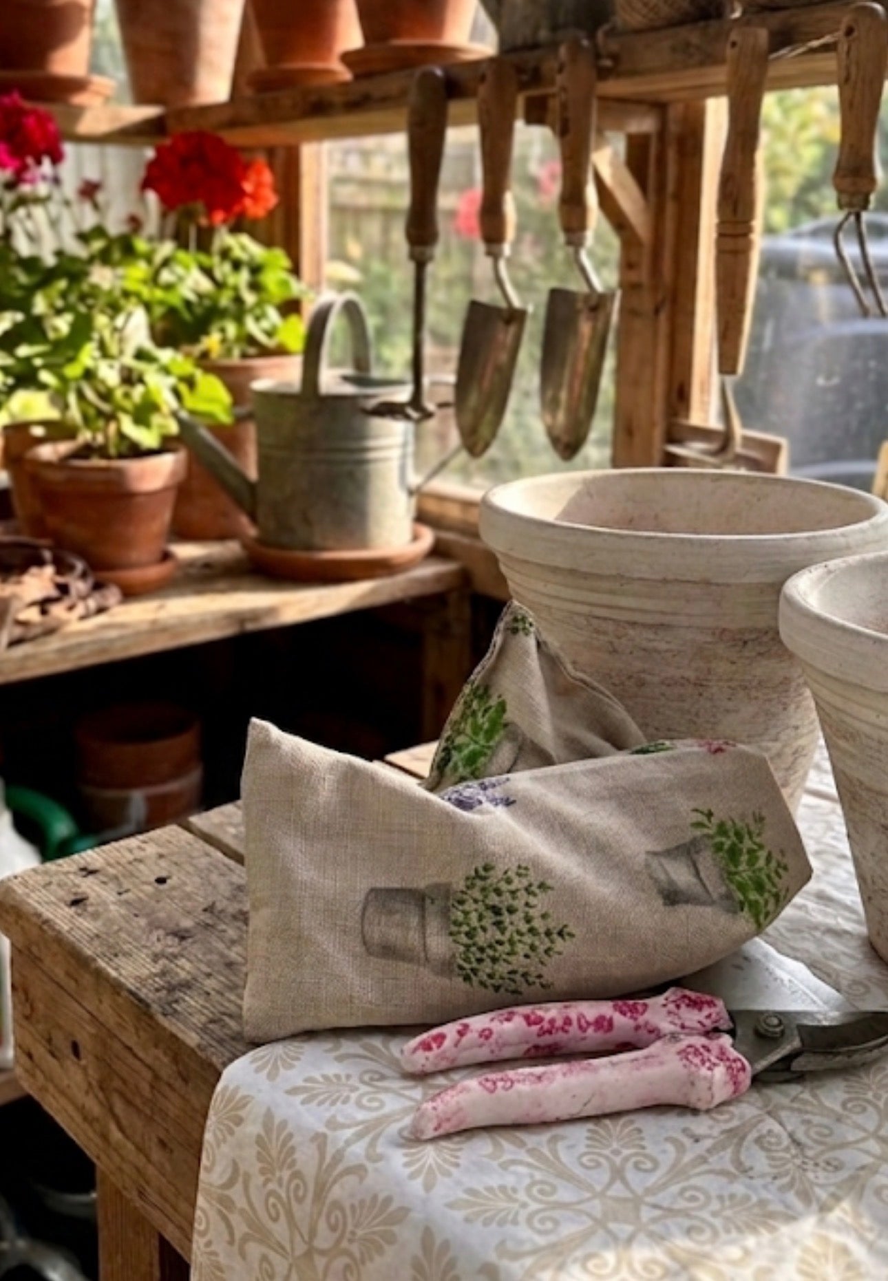 Garden potting shed with geraniums in terracotta pots on shelf and a wooden bench with white clay pots, secateurs and a lavender wheat bag. On a sunny bright day, the gardeners retreat. 