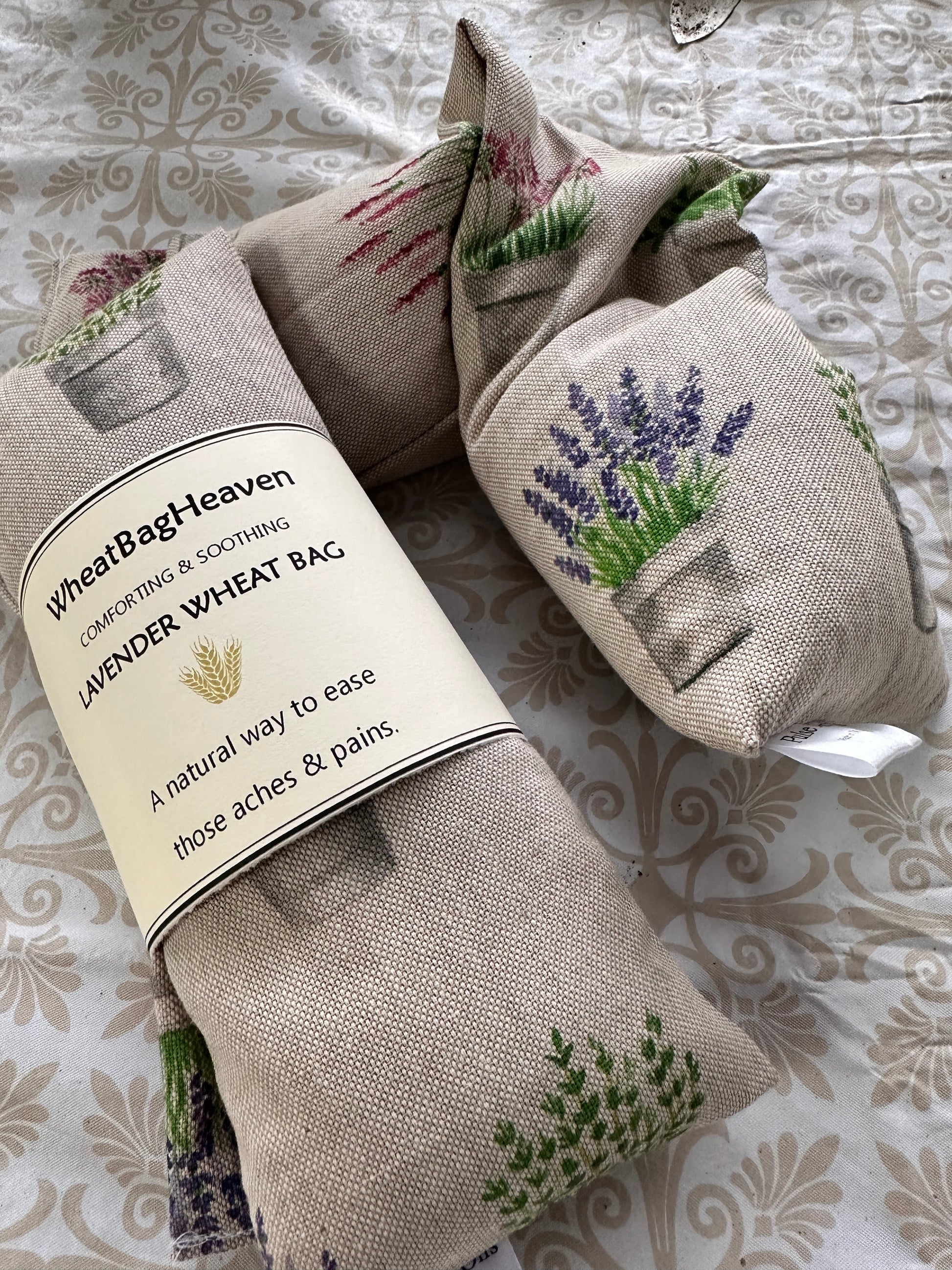 Lavender wheat bags with an all over pot plant print including alliums, lavender, salvia, basil on a beige base. The label reads WheatBagHeaven comforting and soothing Lavender Wheat Bag a natural way to ease those aches and pains 
