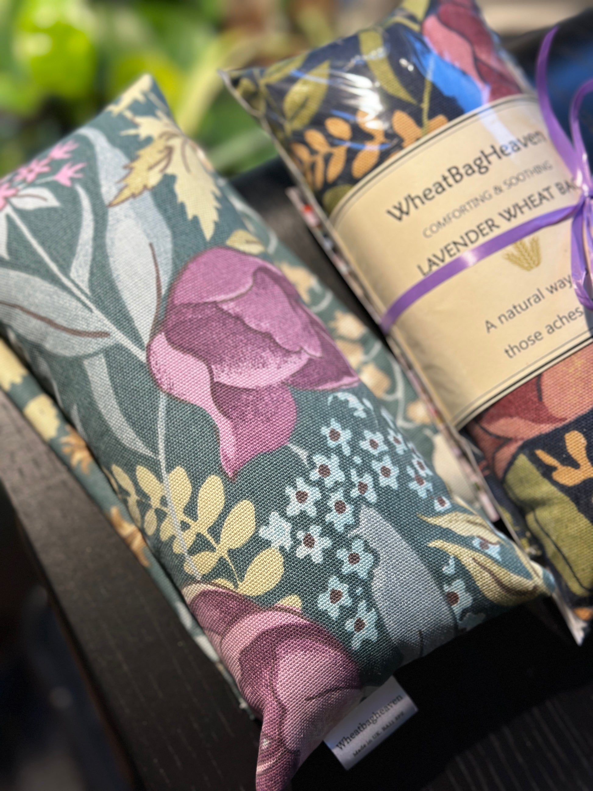Two Lavender Wheat bags from WheatBagHeaven  both with floral design, the left is called Cottage Garden in shades of pink, blue and green on a darker sage green base the right is called Midnight Garden same flowers in golds, pinks and green on a navy base with label that reads WheatBagHeaven, comforting and soothing Lavender Wheat Bag. A natural way to ease those aches and pains also makers tag. 