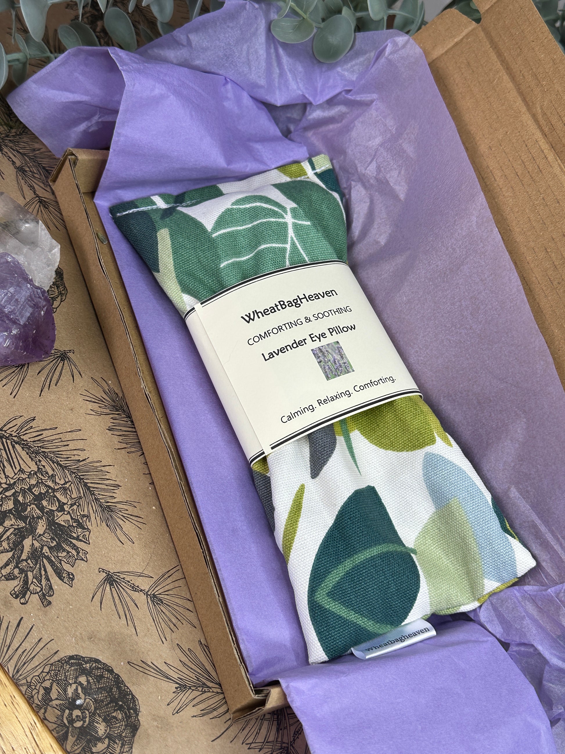 Packaged eye pillow with an all over leaf pattern. The eye pillow is laid in a box on purple tissue paper with maker's tag and belly band that reads WheatBagHeaven, comforting and soothing lavender eye pillow with a picture of lavender in a field under text