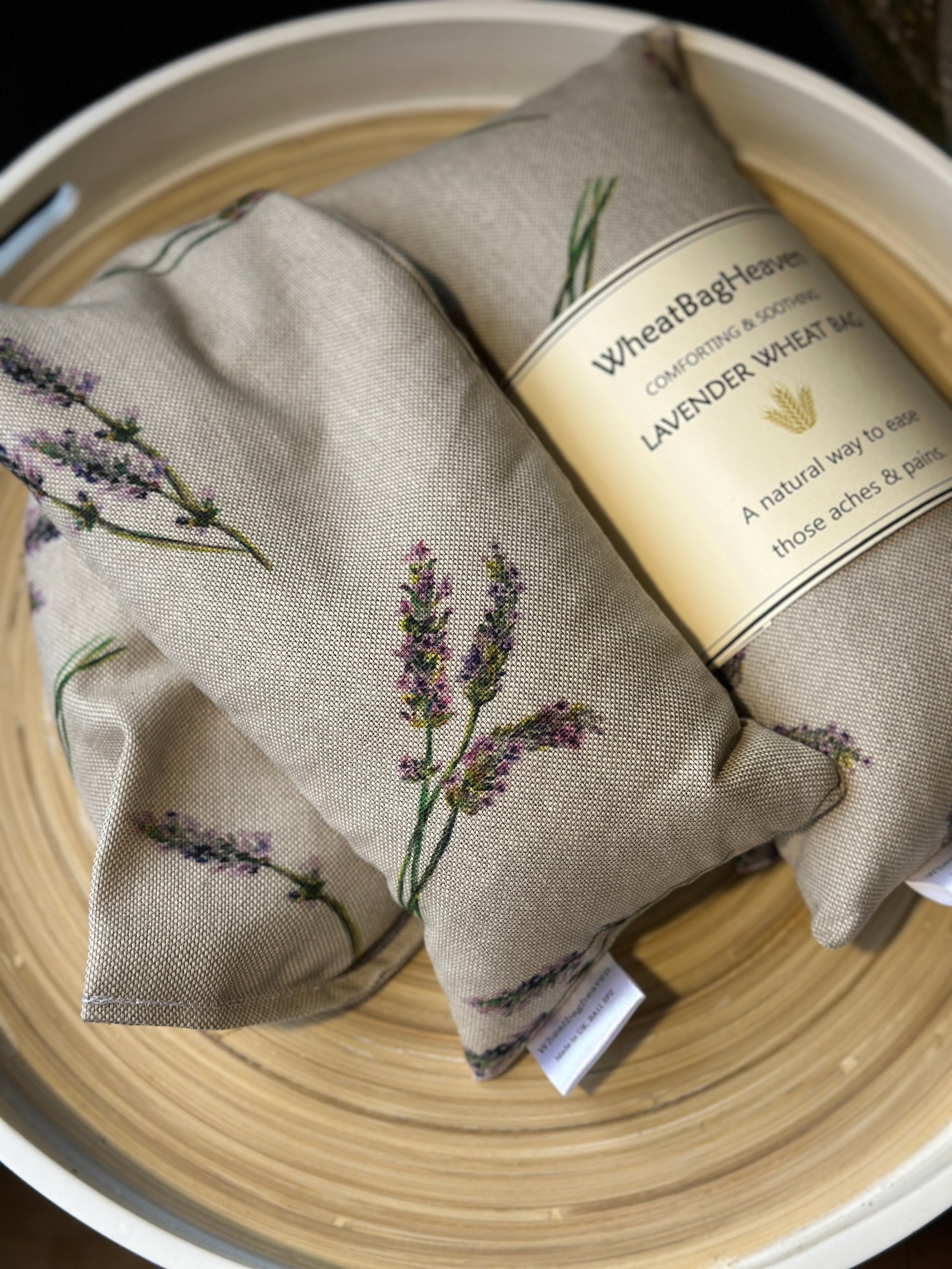 closer look at Two Wheat bags with a lavender flower print design on a wooden surface. One wheat bag has maker's belly band that read WheatBagHeaven comforting and soothing Lavender Wheat Bag A natural way to ease those aches and pains. Both have makers tags