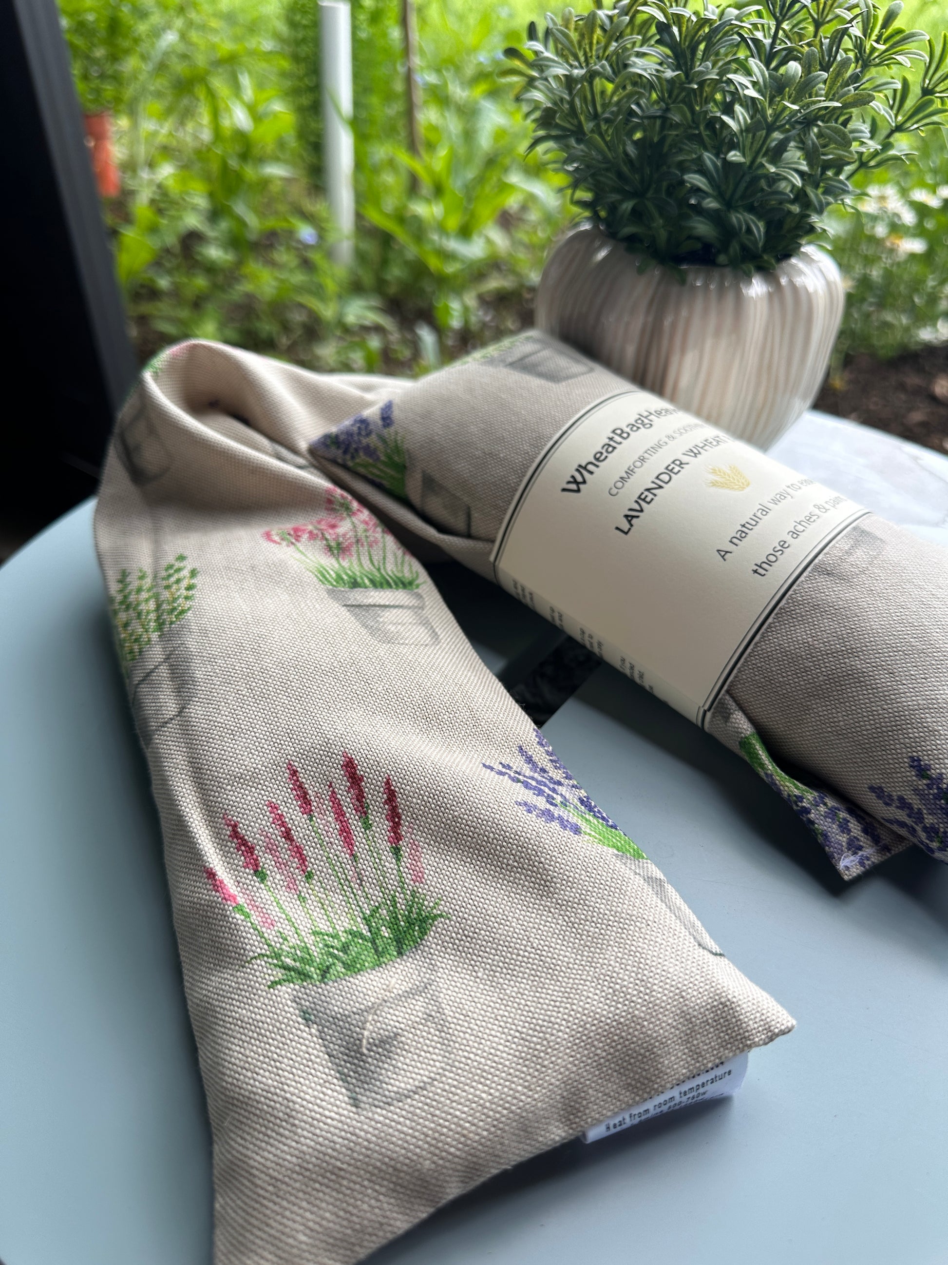Lavender wheat bags with an all over pot plant print including alliums, lavender, salvia, basil on a beige base. The label reads WheatBagHeaven comforting and soothing Lavender Wheat Bag a natural way to ease those aches and pains 