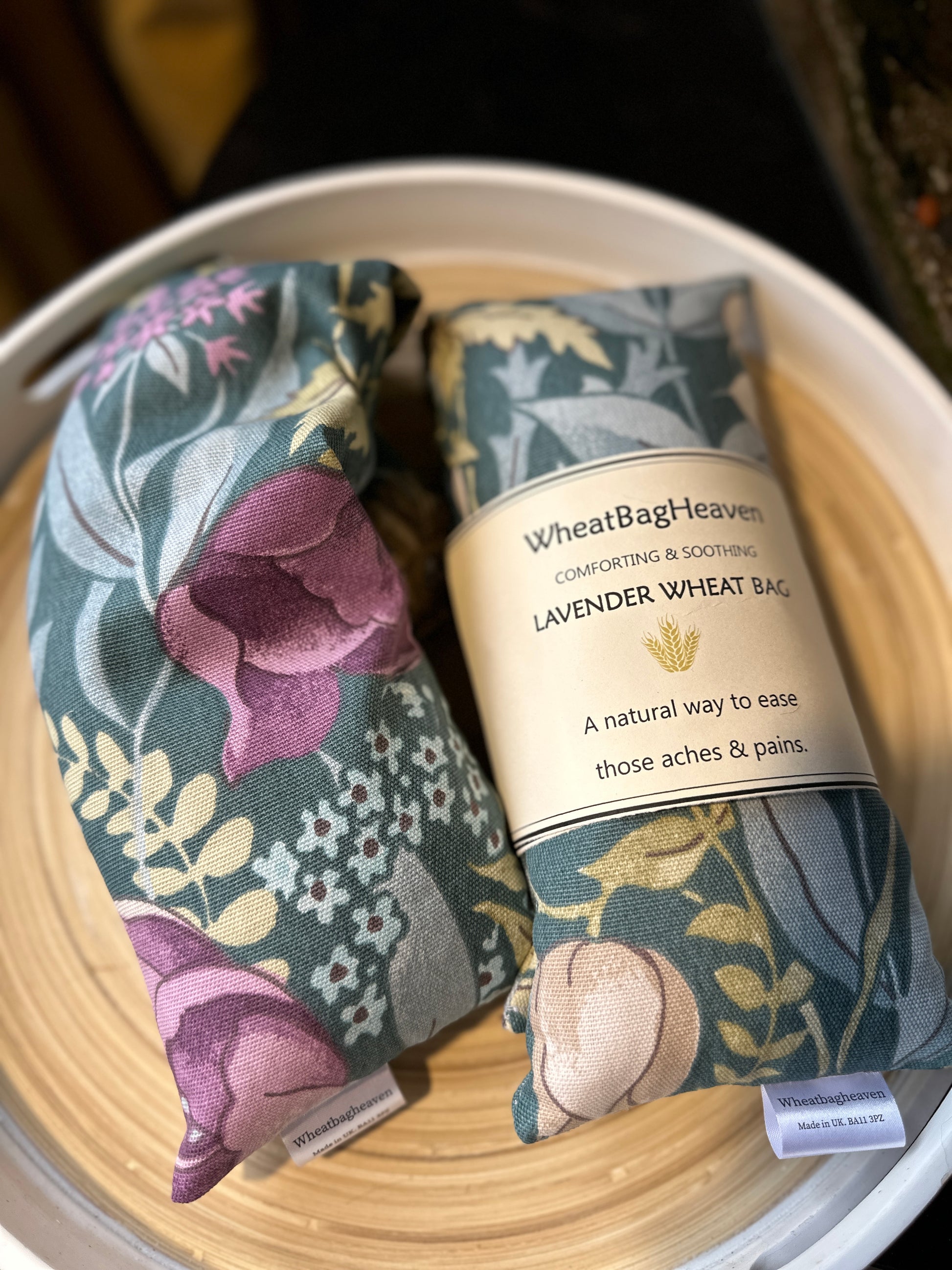 Two Lavender Wheat bags from WheatBagHeaven with floral design called Cottage Garden in shades of pink, blue and green on a darker sage green base with label that reads WheatBagHeaven, comforting and soothing Lavender Wheat Bag. A natural way to ease those aches and pains also makers tag. The wheat bags are laid on a wooden tray