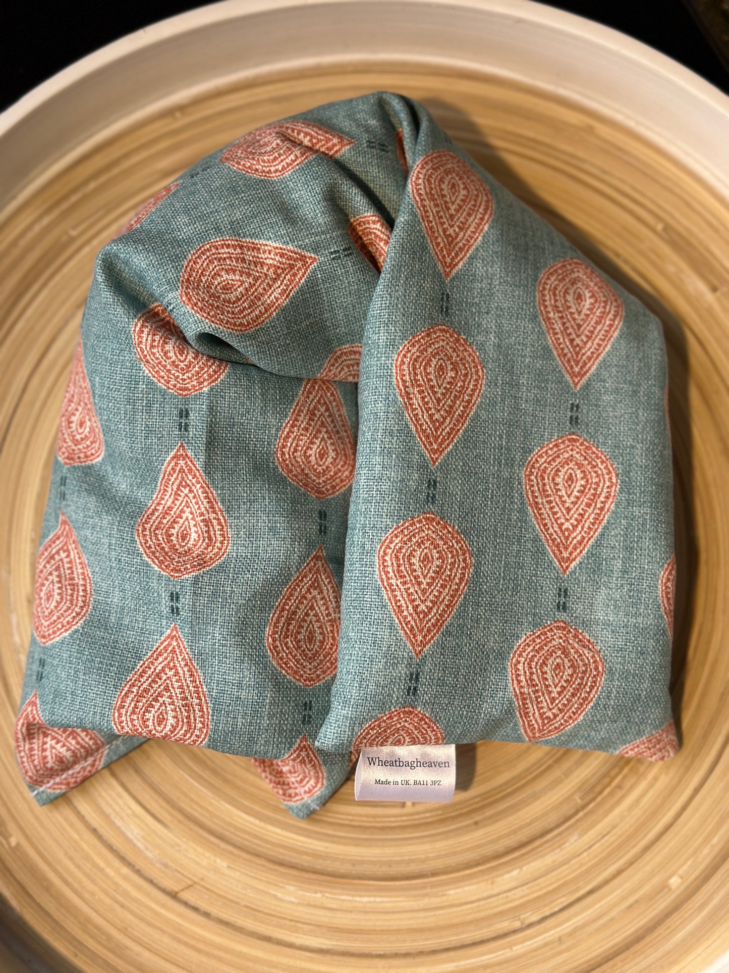 An unlabelled lavender wheat bag on a wooden tray. The wheat bag has a paisley teardrop pattern in shades of pink and cream with a base colour of teal. With a maker's tag that reads WheatBagHeaven