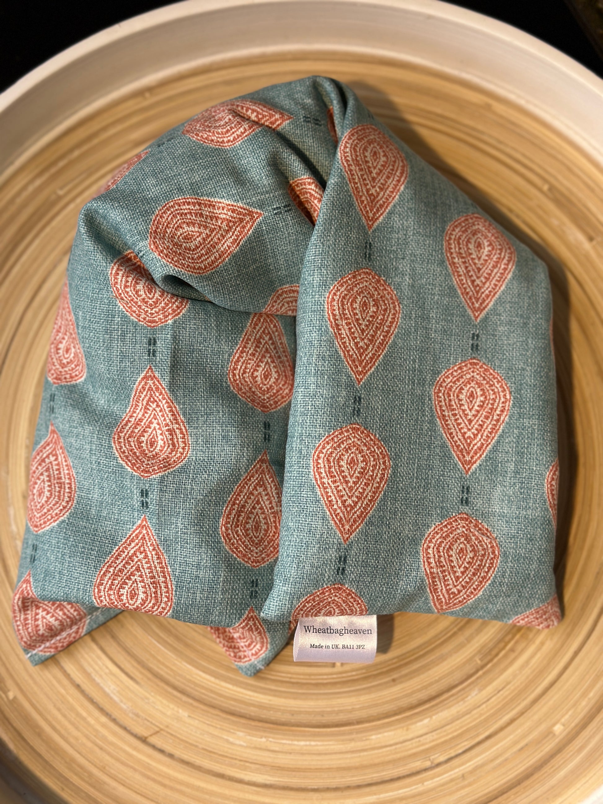 An unlabelled lavender wheat bag on a wooden tray. The wheat bag has a paisley teardrop pattern in shades of pink and cream with a base colour of teal. With a maker's tag that reads WheatBagHeaven