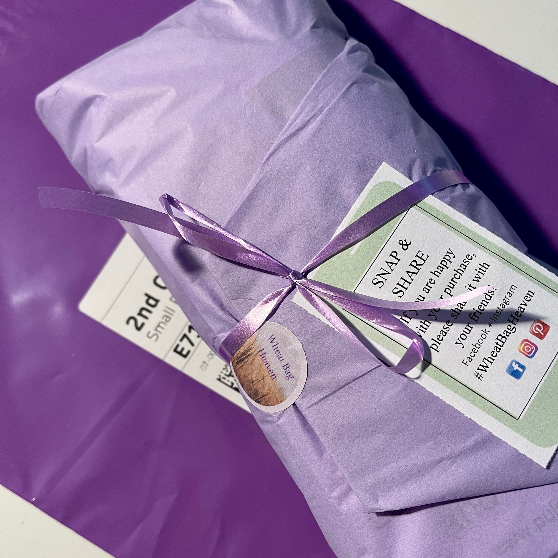 Wheat bag wrapped in lilac tissue paper tied with purple ribbon with a call to action card. Wheat bag is sat on a purple recyclable postal bag