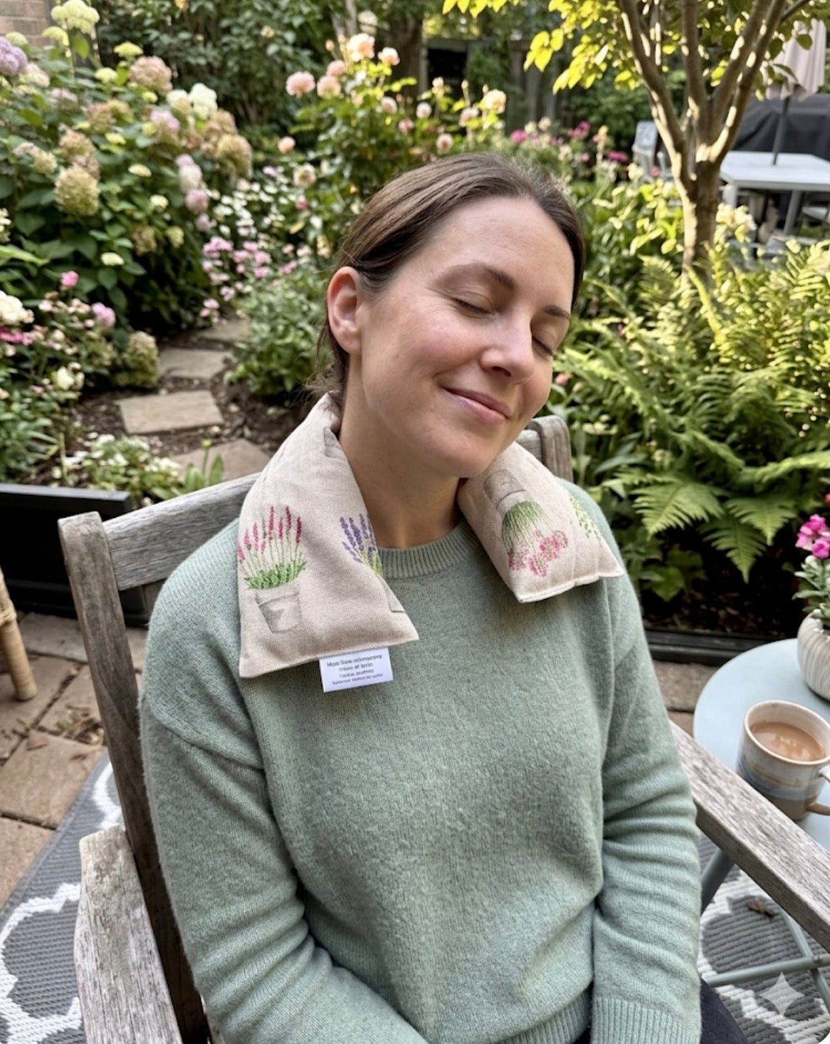 Woman sitting relaxing outdoors with a floral-patterned lavender wheat bag used as a neck wrap, she is surrounded by garden plants.