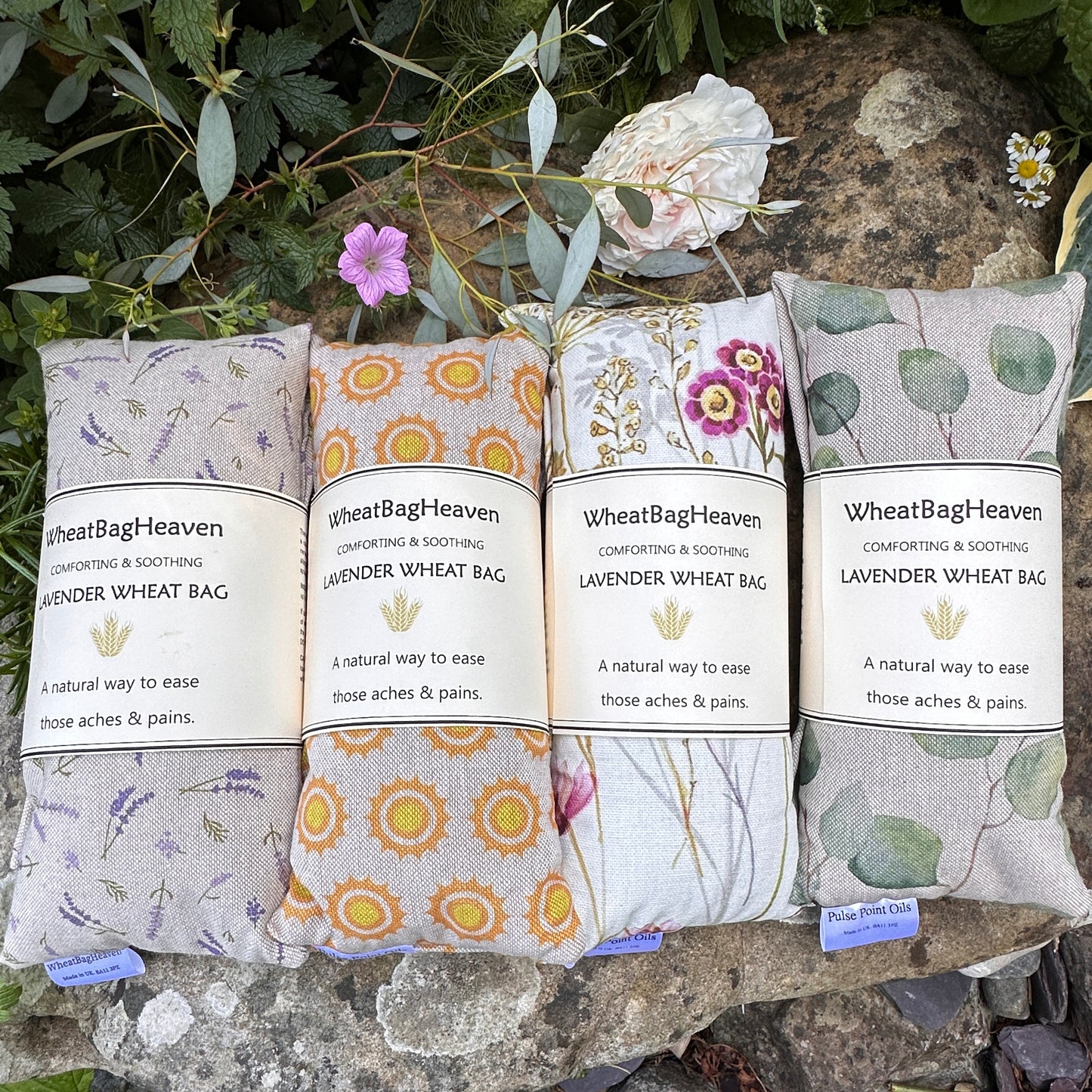 Four in a row, lavender scented wheat bags from wheat bag heaven from left to right we have small lavender sprigs print, then sunshine, wetlands floral print and finally eucalyptus leaf, side by side on a stone backdrop in the gardens of WheatBagHeaven.com