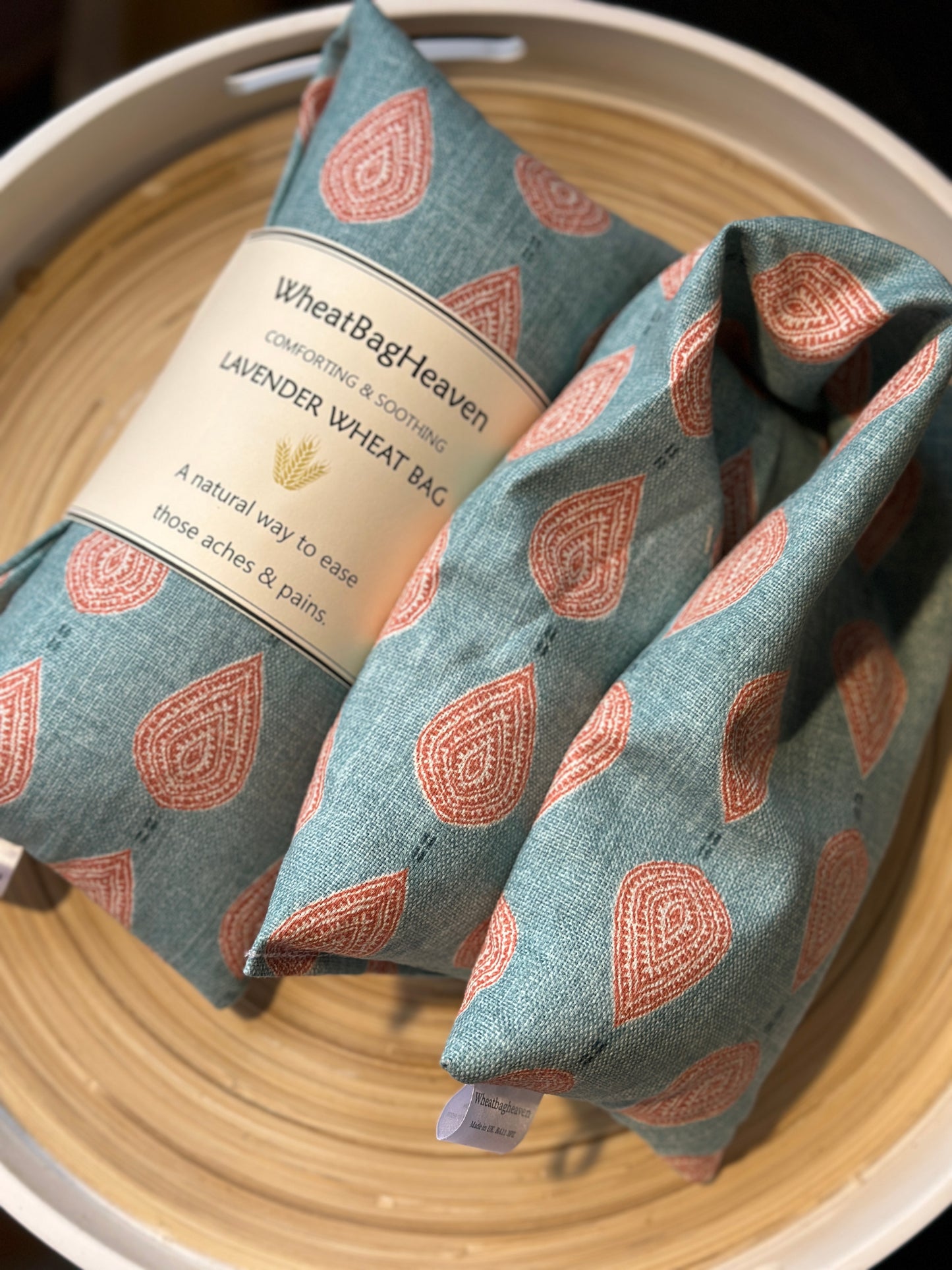 Two lavender wheat bags on a wooden tray. The wheat bags have a paisley teardrop pattern in shades of pink and cream with a base colour of soft teal. Its labelled "WheatBagHeaven" comforting and soothing lavender wheat bag. A natural way to ease those aches and pains. Also a tag that reads WheatBagHeaven