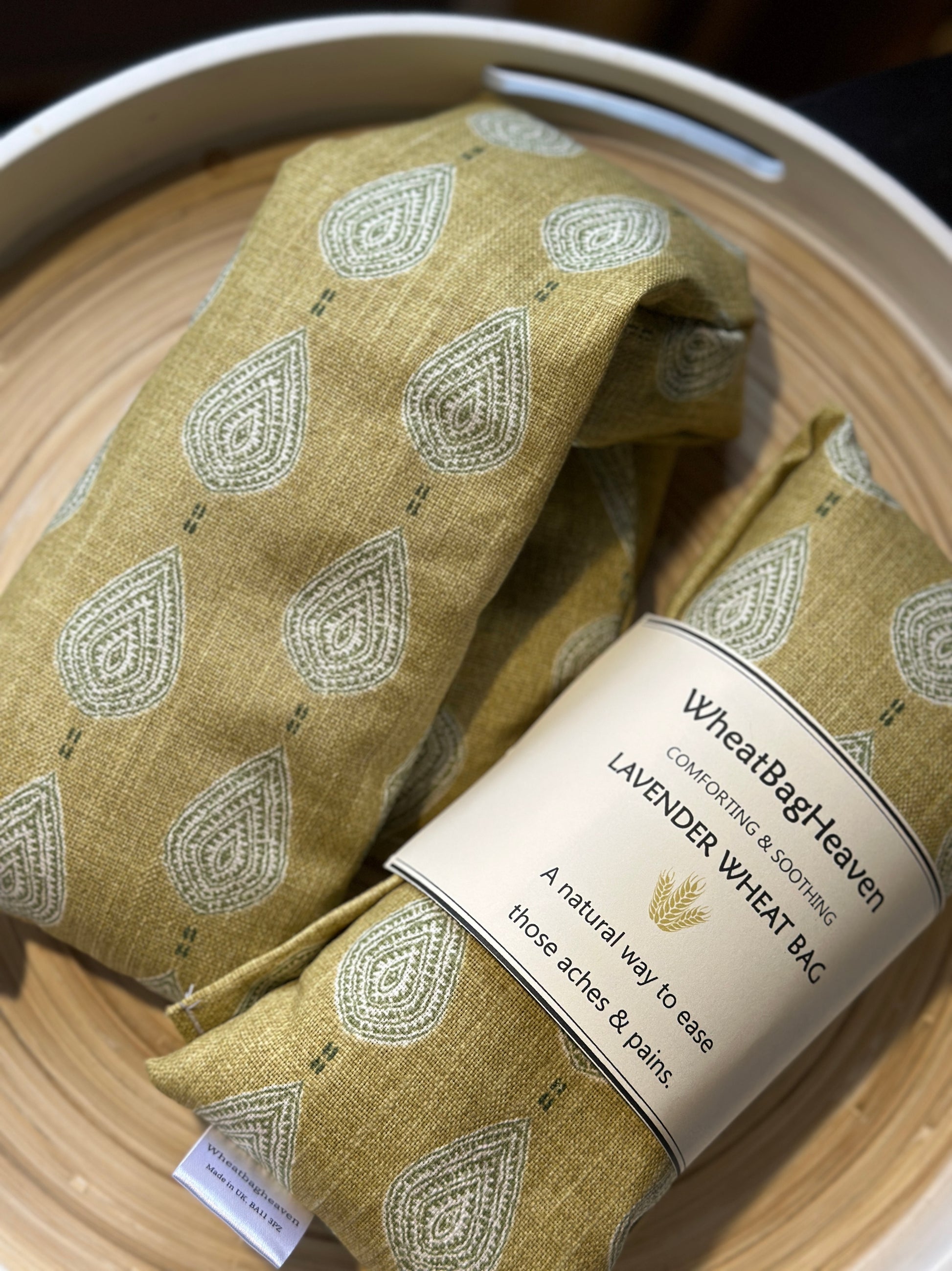 A lavender wheat bag on a wooden tray. The wheat bag has a paisley teardrop pattern in shades of green and cream with a base colour of ochre. Its labelled "WheatBagHeaven" comforting and soothing lavender wheat bag. A natural way to ease those aches and pains. Also a tag that reads WheatBagHeaven