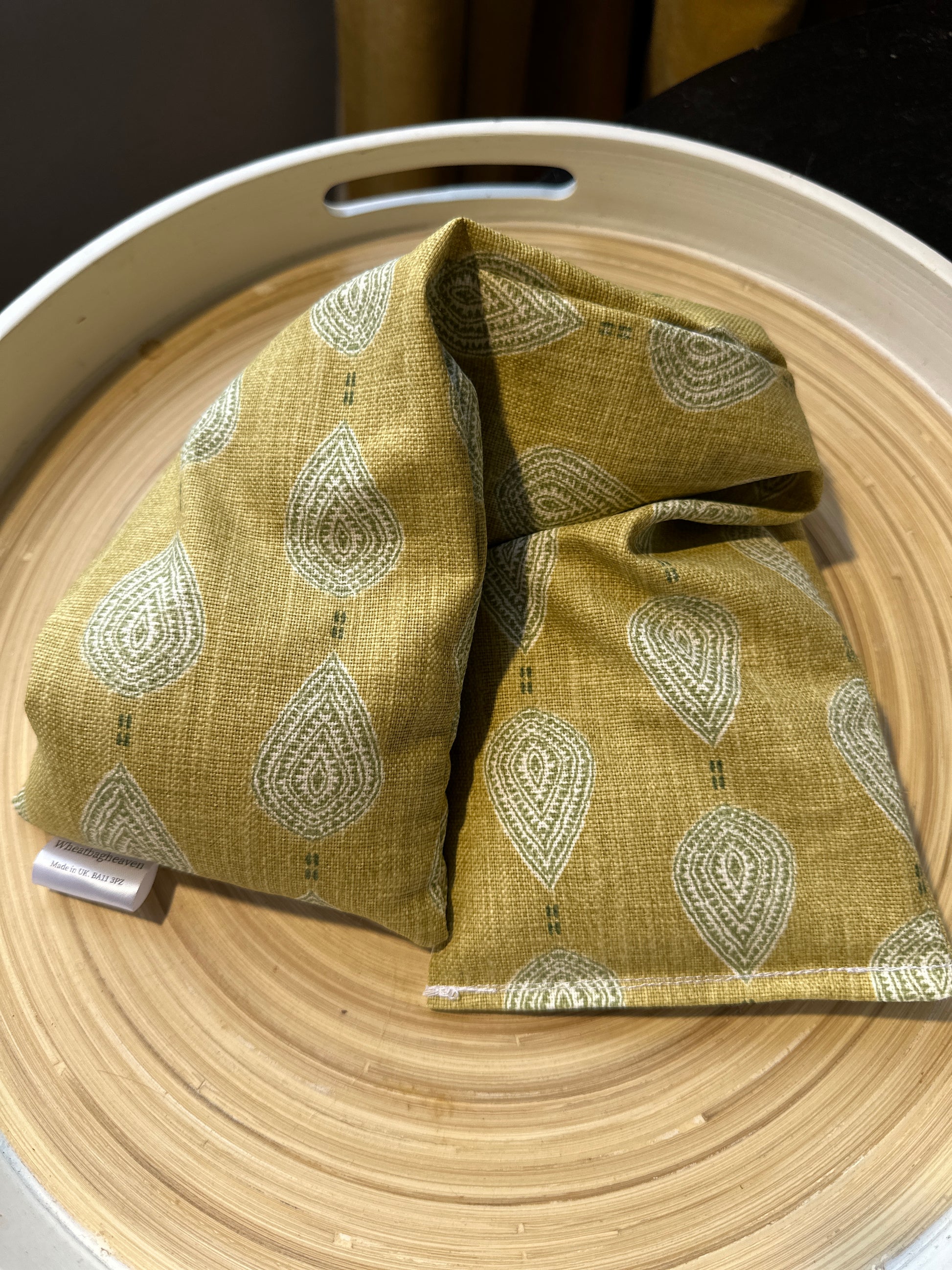 close up picture of a lavender wheat bag on a wooden tray. The wheat bag has a paisley teardrop pattern in shades of green and cream with a base colour of ochre with makers tag that reads WheatBagHeaven