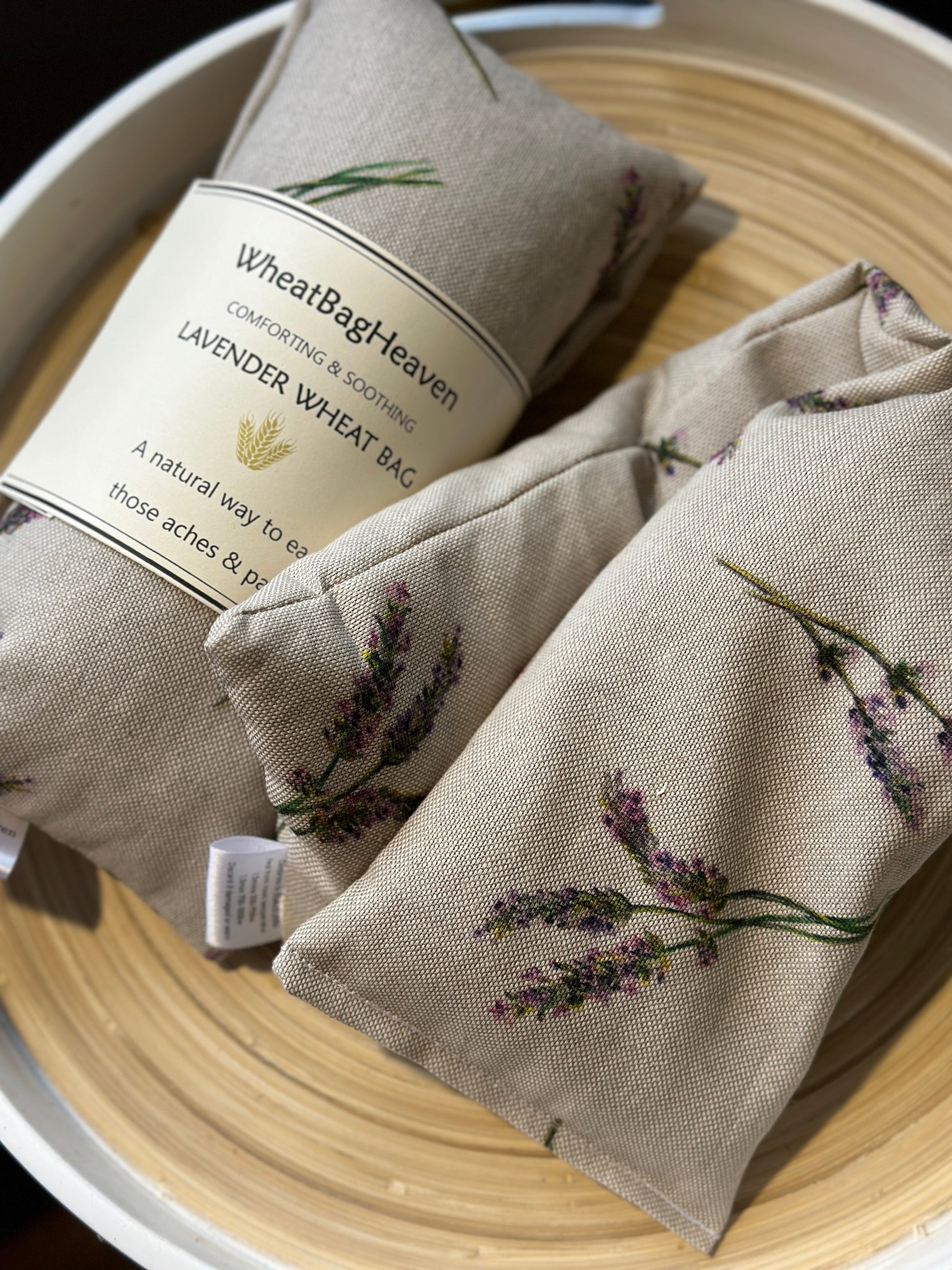 Two Wheat bags with a lavender sprigs print design on a wooden surface. One wheat bag has maker's belly band that read WheatBagHeaven comforting and soothing Lavender Wheat Bag A natural way to ease those aches and pains. Both have makers tags