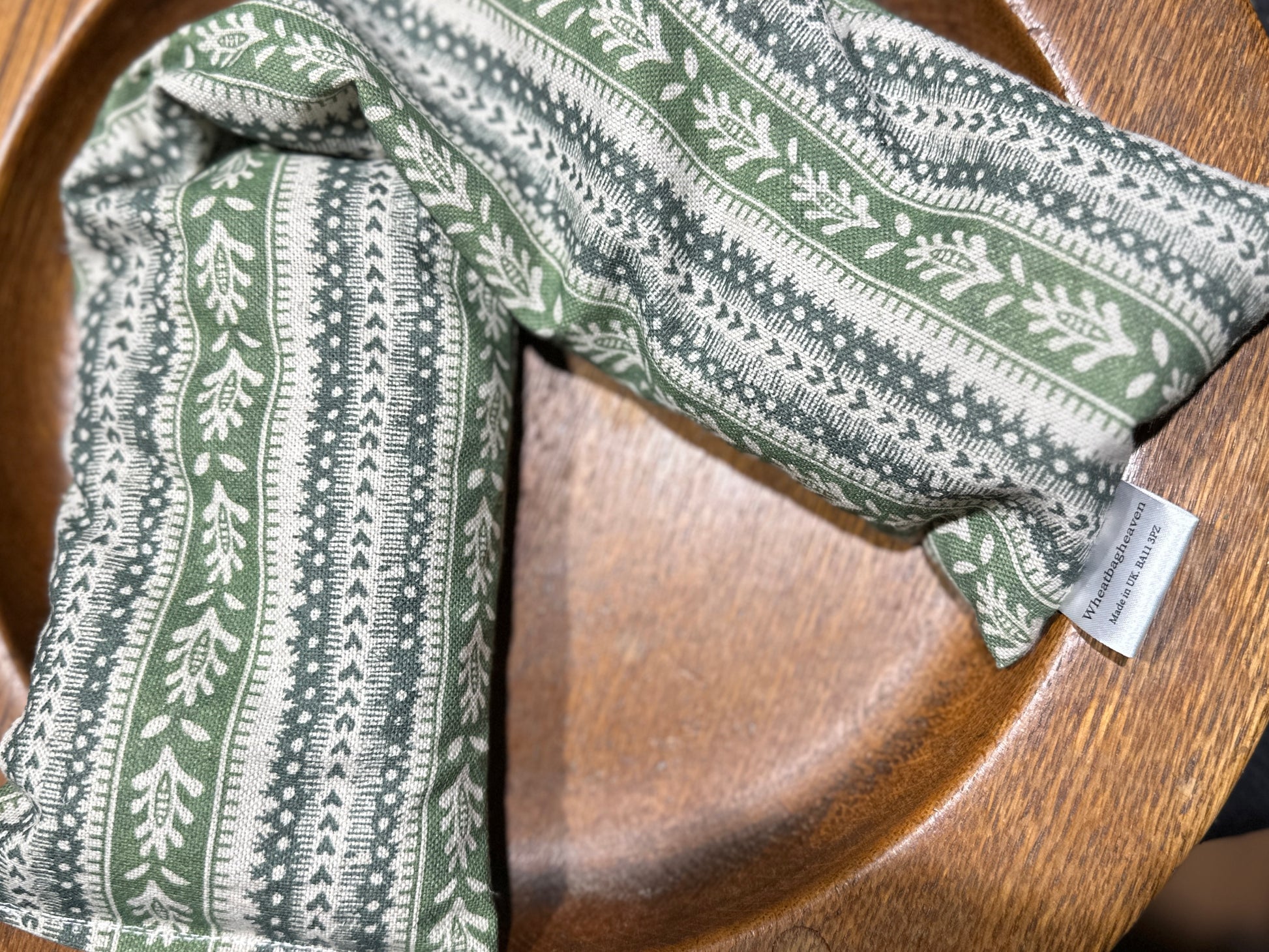A lavender scented wheat bag with a green heart stripe and botanical pattern on a wooden surface. Has a maker's tag that reads WheatBagHeaven