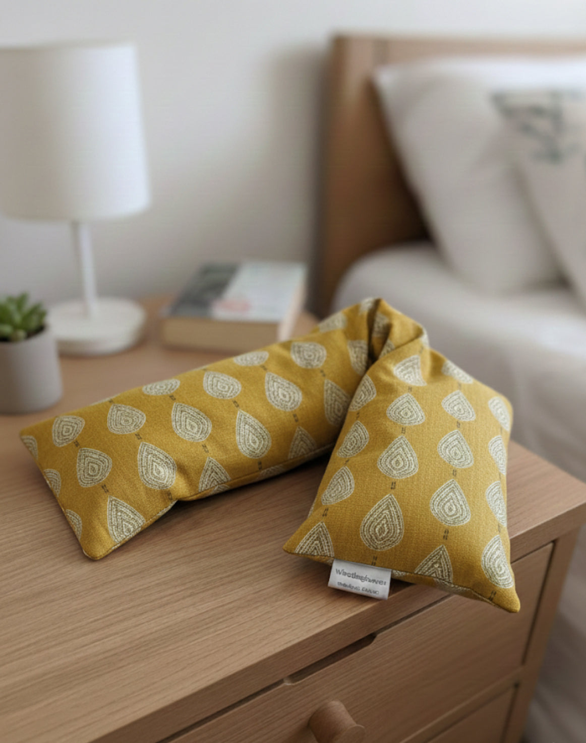 A wheat bag with a paisley teardrop pattern in shades of green and cream with a base colour of ochre and makers tag that reads WheatBagHeaven. The wheat bag is sat on a wooden bedside table next to a book, plant pot and lamp with a bed in the background