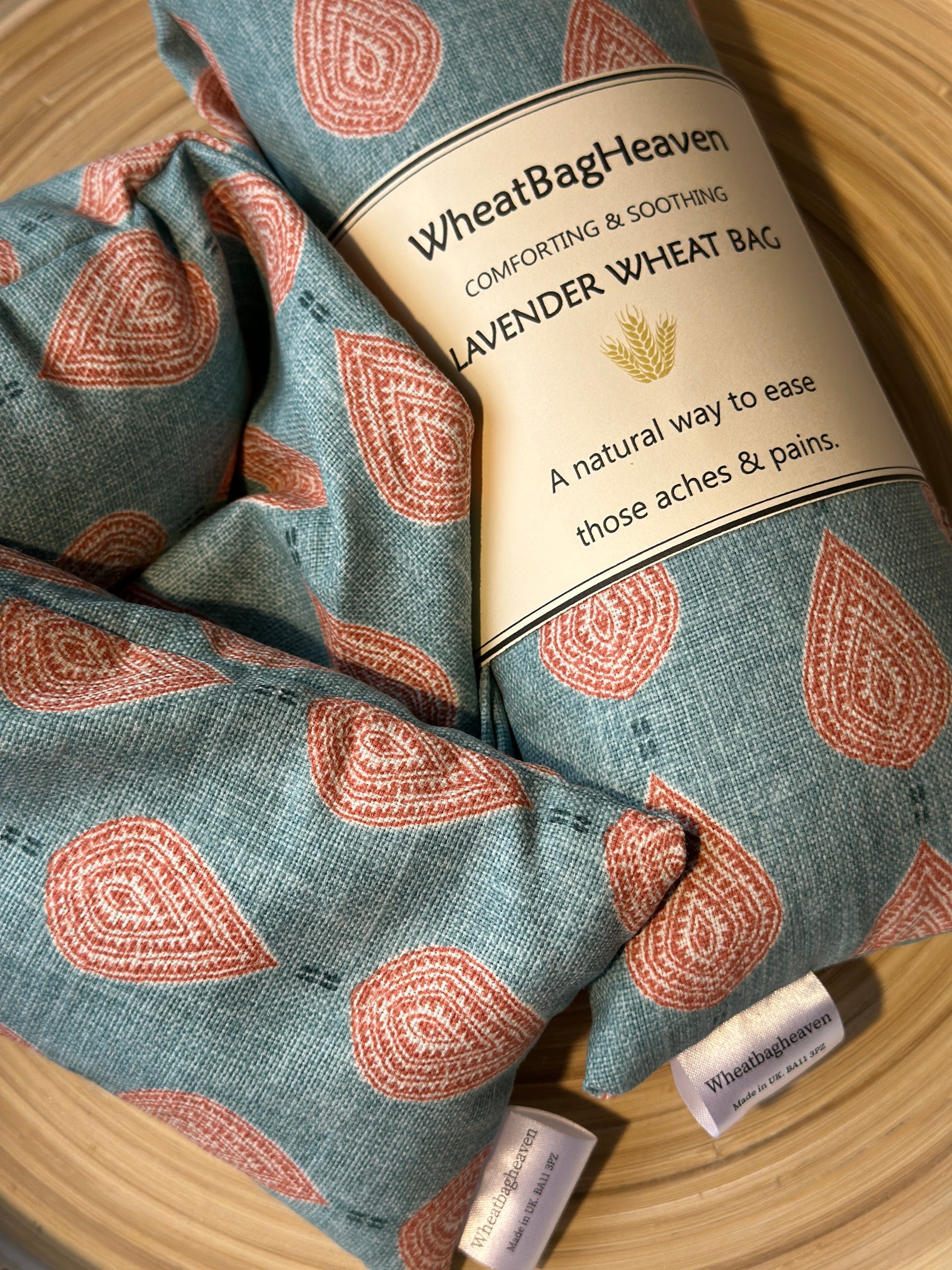 A lavender wheat bag on a wooden tray. The wheat bag has a paisley teardrop pattern in shades of pink and cream with a base colour of teal. Its labelled "WheatBagHeaven" comforting and soothing lavender wheat bag. A natural way to ease those aches and pains. Also a tag that reads WheatBagHeaven