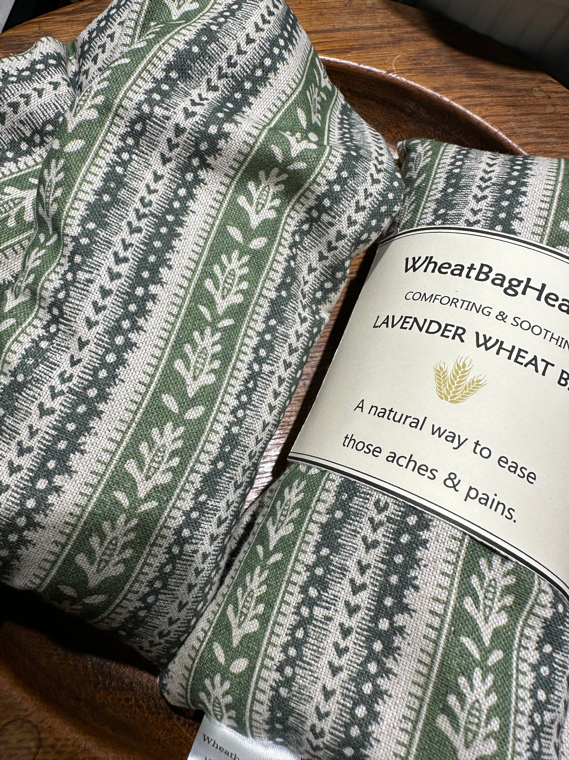 Two lavender scented wheat bags with a green heart stripe and botanical pattern on a wooden surface. One is labelled WheatBagHeaven comforting and soothing lavender wheat bag, a natural way to ease those aches and pains.