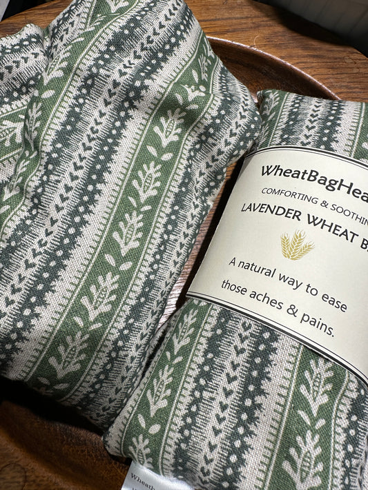 Two lavender scented wheat bags with a green heart stripe and botanical pattern on a wooden surface. One is labelled WheatBagHeaven comforting and soothing lavender wheat bag, a natural way to ease those aches and pains.