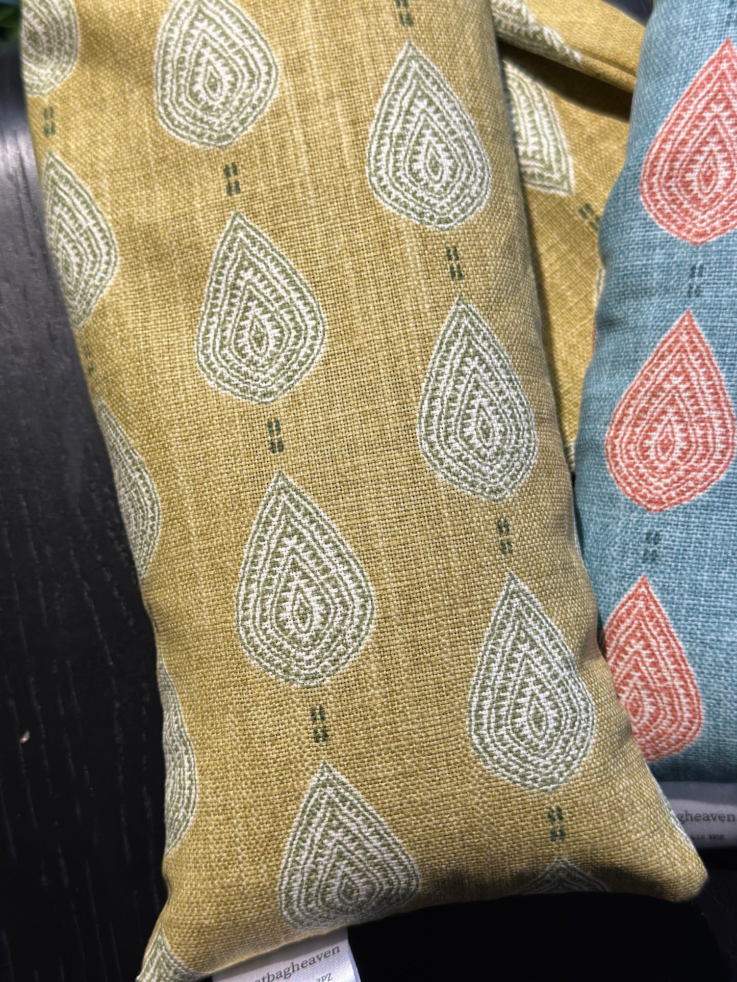 close up of the wheat bag fabric paisley teardrop pattern in shades of green and cream with a base colour of ochre. The wheat bag has a tag that reads WheatBagHeaven