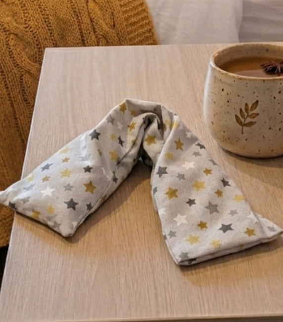 Close up of Yellow star lavender wheat bag from WheatBagHeaven. The stars are in colours of ochre, greys, white on a beige base and sat on bedside table next to a mug of coffee, in the background is a bed with ochre knitted throw 