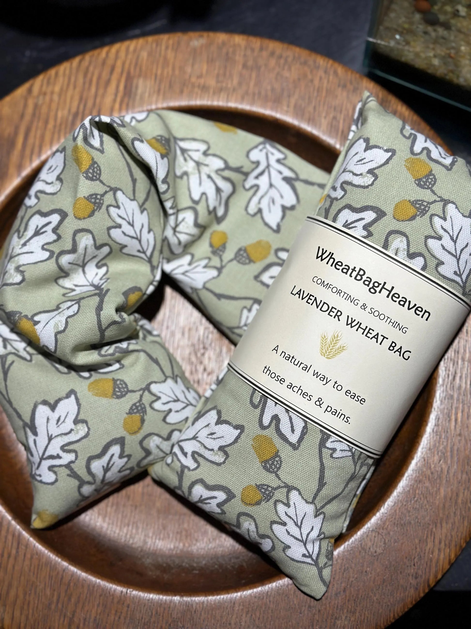 acorn wood wheat bag from WheatBagHeaven in sage green base with an all over print of white leaves outlined in grey with brown and ochre acorns. The label reads 'WheatBagHeaven' comforting and soothing Lavender Wheat Bag. A natural way to ease those aches and pains