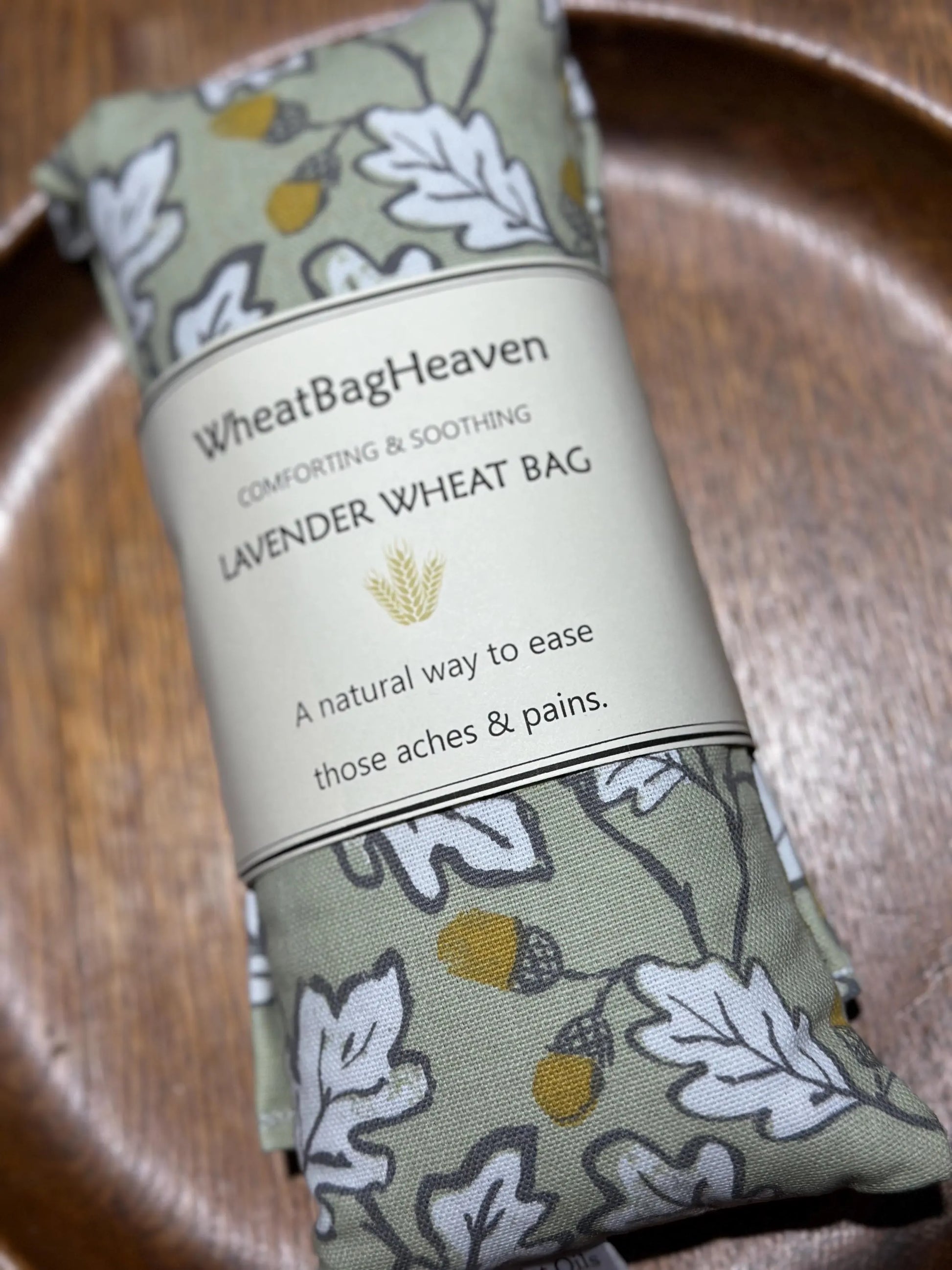 acorn wood wheat bag from WheatBagHeaven on a wooden bowl, The wheat bag is sage green with an all over print of white leaves outlined in grey with brown and ochre acorns. The label reads 'WheatBagHeaven' comforting and soothing Lavender Wheat Bag. A natural way to ease those aches and pains. Perfect gardeners gift