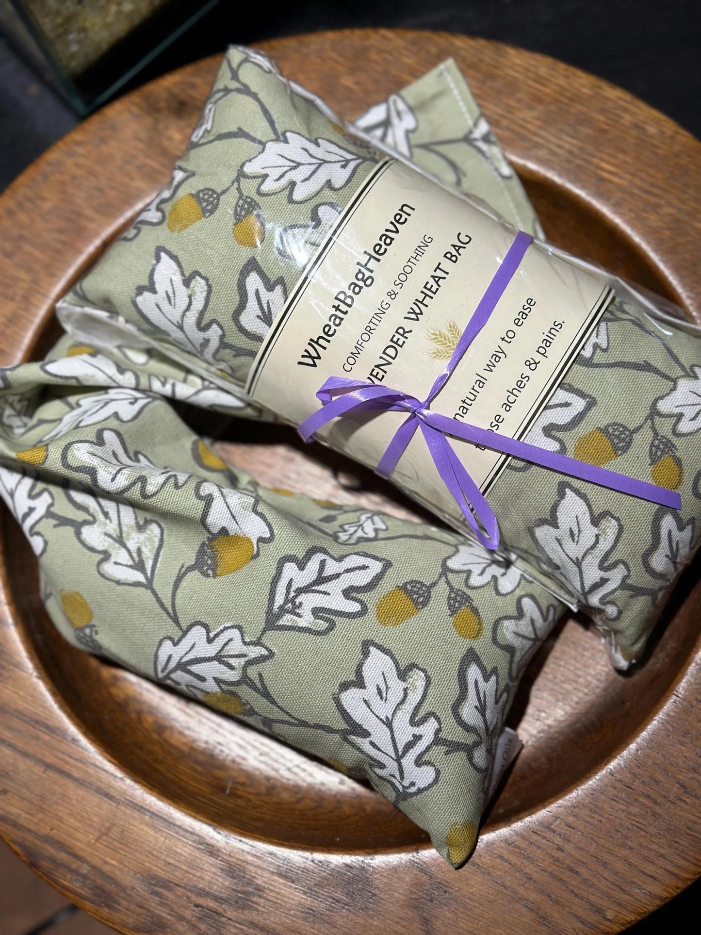 acorn wood wheat bags from WheatBagHeaven with a sage green base  and all over print of white leaves outlined in grey with brown and ochre acorns. The label reads 'WheatBagHeaven' comforting and soothing Lavender Wheat Bag. A natural way to ease those aches and pains and is tied with purple ribbon
