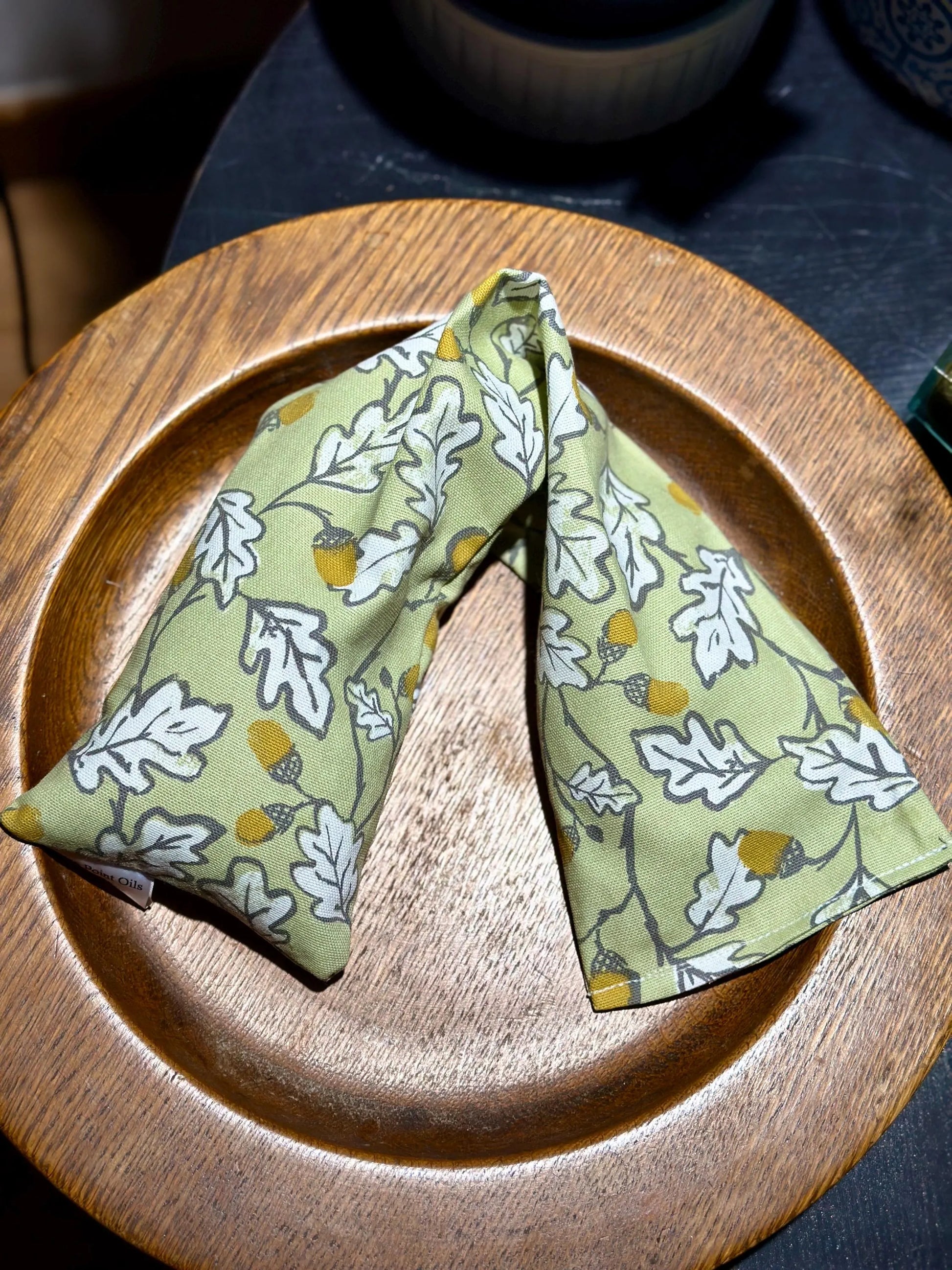 In a wooden bowl sits an acorn wood  printed wheat bag from WheatBagHeaven. Sage green base with an all over print of white leaves outlined in grey with brown and ochre acorns.  Warming neck heat wrap