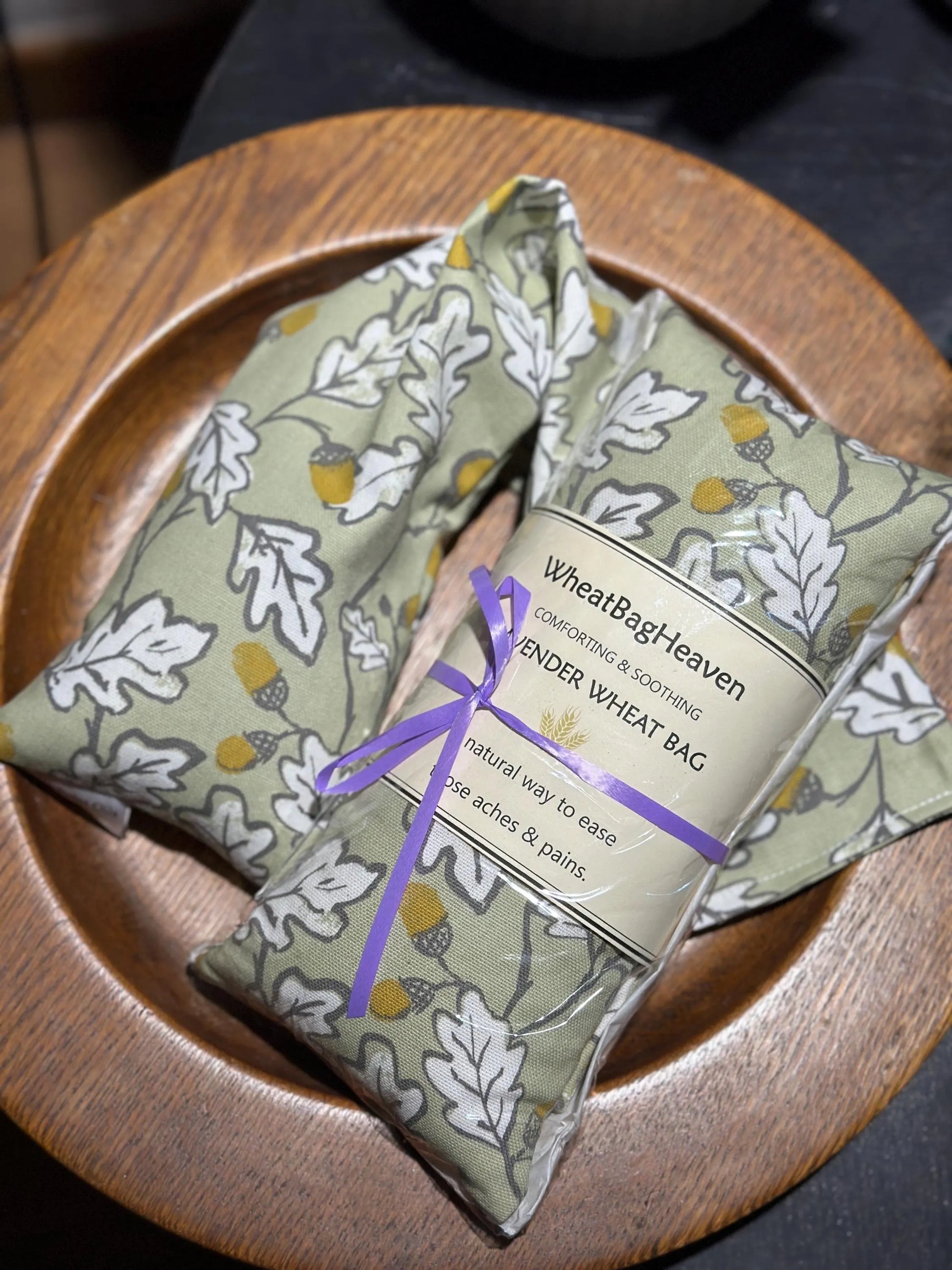 Two acorn wood wheat bags from WheatBagHeaven in sage green base with an all over print of white leaves outlined in grey with brown and ochre acorns. The label reads 'WheatBagHeaven' comforting and soothing Lavender Wheat Bag. A natural way to ease those aches and pains. Gardener's heating pad to soothe those aching backs