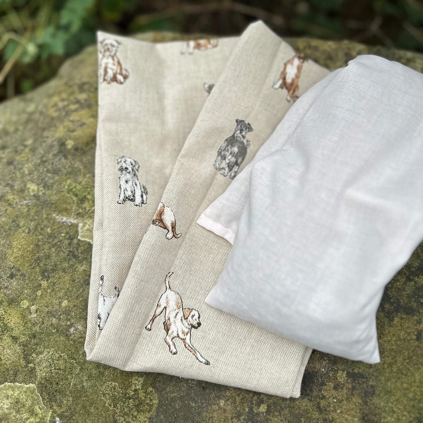 Folded wheat bag washable cover with an all over dog print, Terrier, Boxer, Labrador, Spaniel, Scottie. In shades of grey, brown and cream on a beige base. A plain wheat bag is beside cover. The cover has maker's tag WheatBagHeaven and is laid on a stone surface