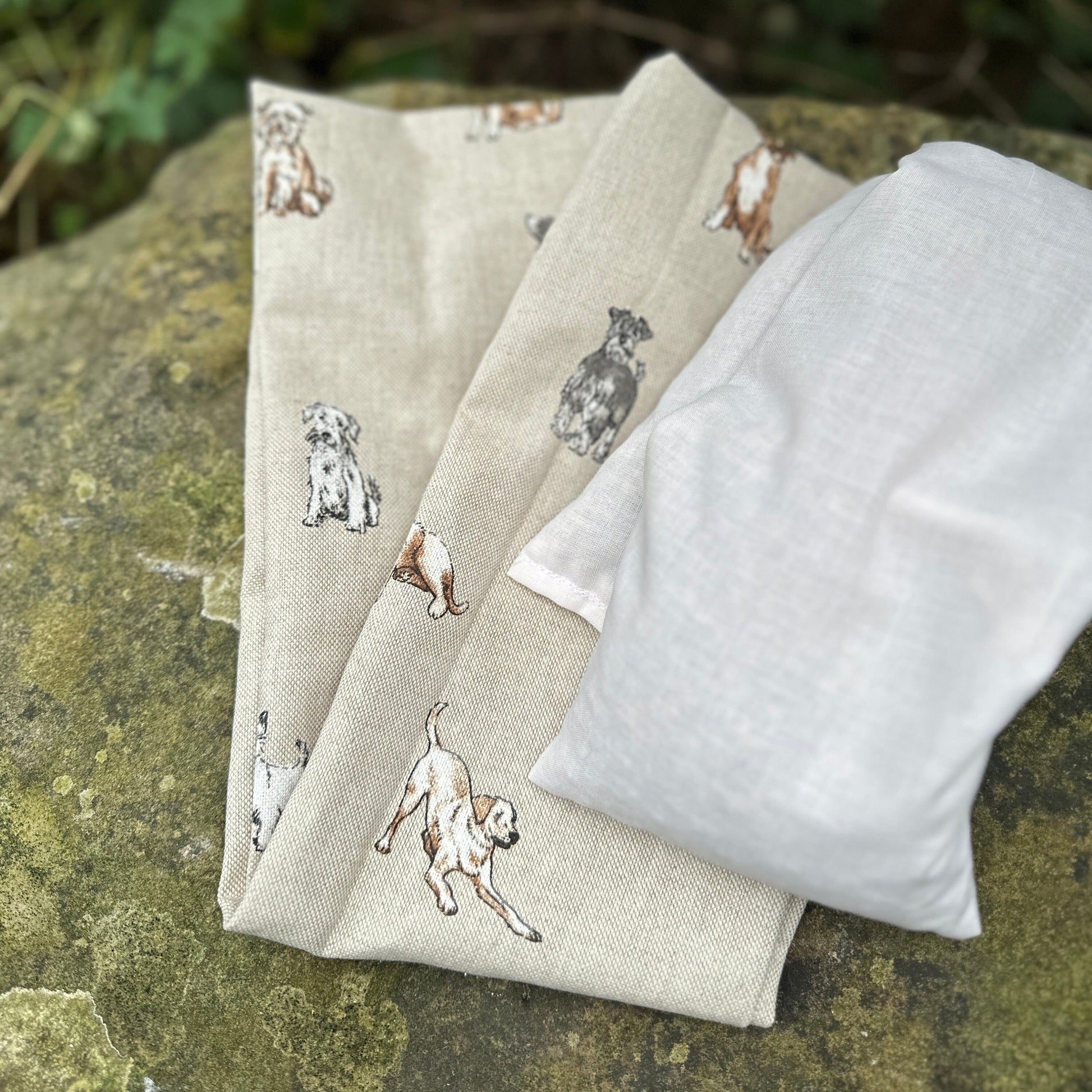 Folded wheat bag washable cover with an all over dog print, Terrier, Boxer, Labrador, Spaniel, Scottie. In shades of grey, brown and cream on a beige base. A plain wheat bag is beside cover. The cover has maker's tag WheatBagHeaven and is laid on a stone surface