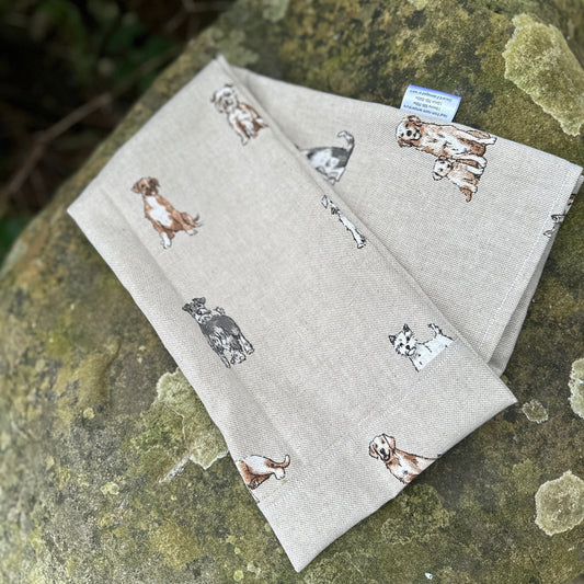 Folded wheat bag washable cover with an all over dog print, Terrier, Boxer, Labrador, Spaniel, Scottie. In shades of grey, brown and cream on a beige base. The cover has maker's tag and is laid on a stone surface