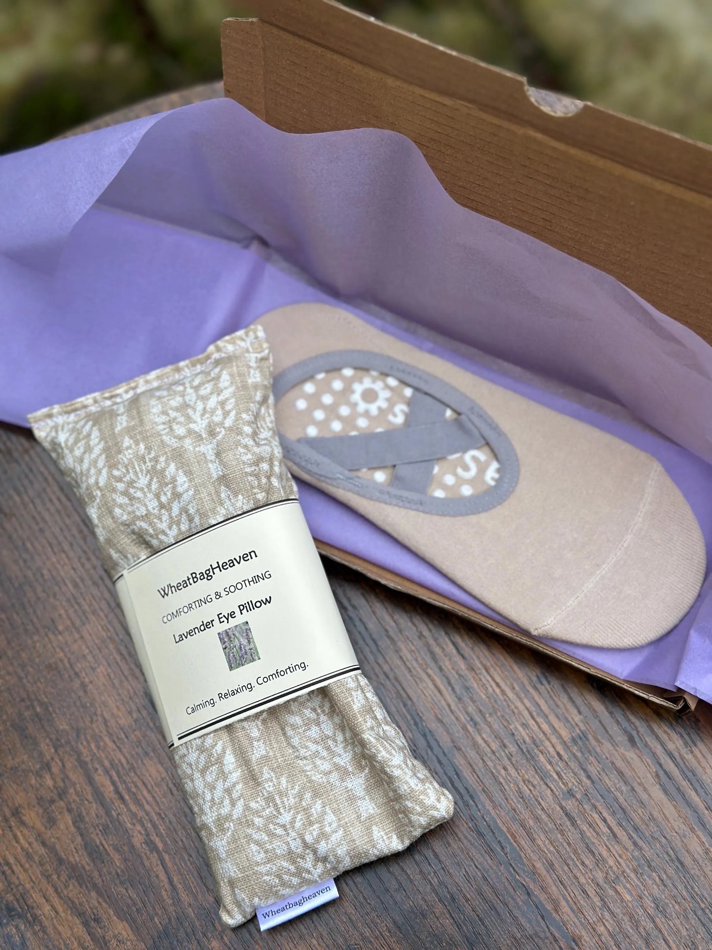 Eye pillow in a cream and beige botanical print with belly band that reads WheatBagHeaven, comforting and soothing Lavender Eye Pillow with makers tag, along side beige yoga socks with lilac packaging on a wooden surface