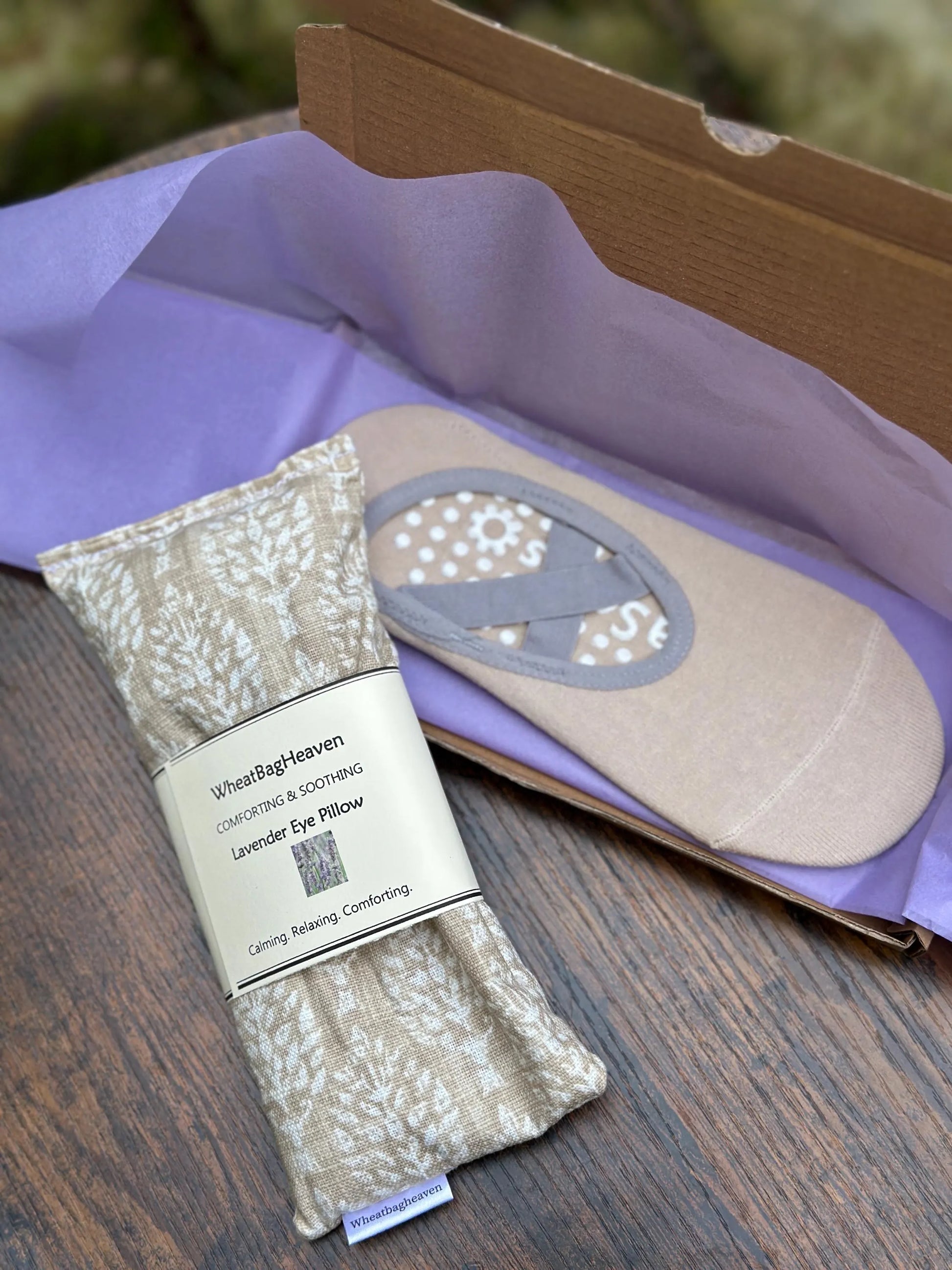 Eye pillow in a cream and beige botanical print with belly band that reads WheatBagHeaven, comforting and soothing Lavender Eye Pillow with makers tag, along side beige yoga socks with lilac packaging on a wooden surface