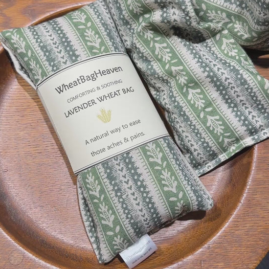 short video of Two lavender scented wheat bags with a green heart stripe and botanical pattern on a wooden surface. One is labelled WheatBagHeaven comforting and soothing lavender wheat bag, a natural way to ease those aches and pains.