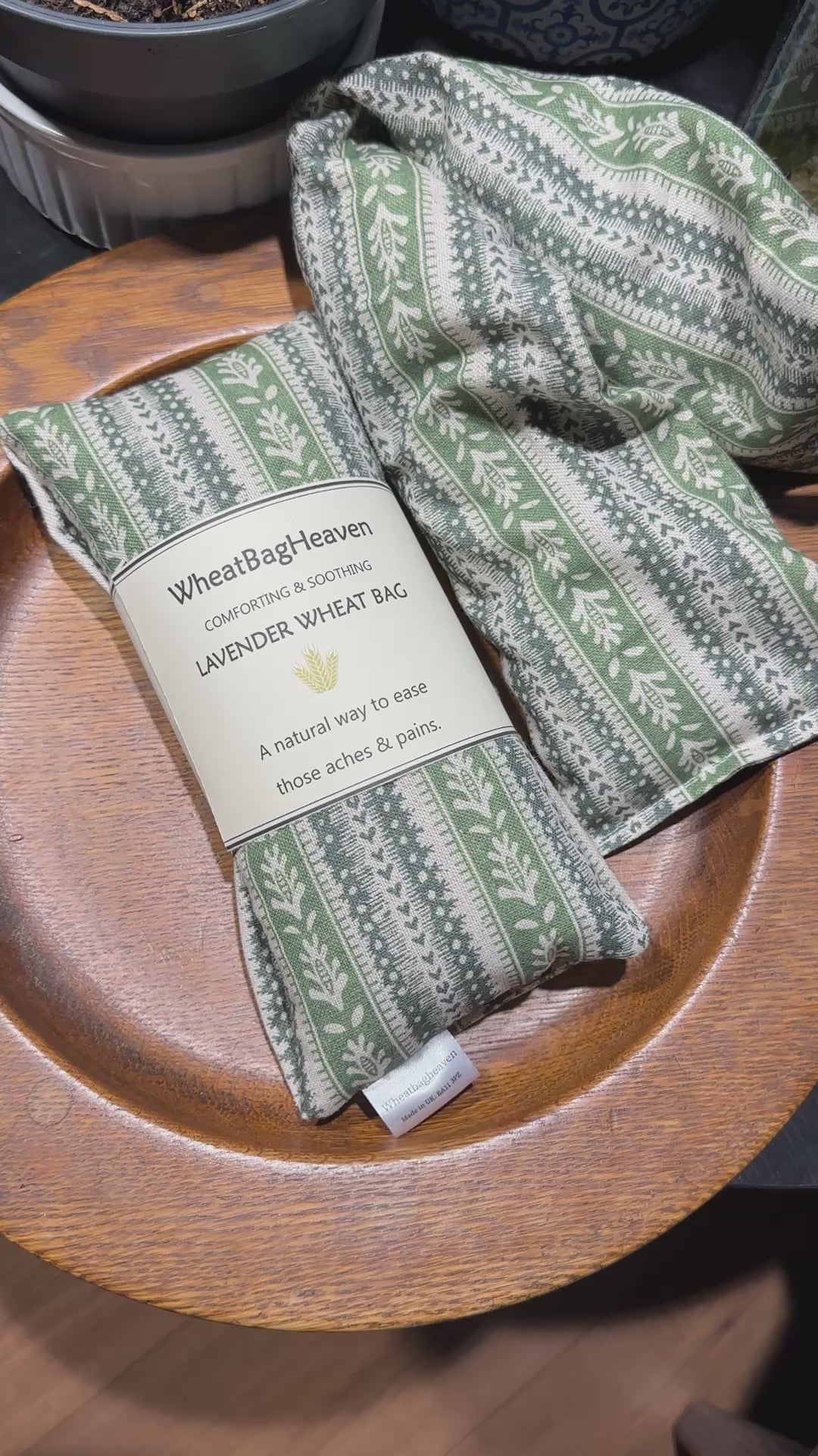short video of Two lavender scented wheat bags with a green heart stripe and botanical pattern on a wooden surface. One is labelled WheatBagHeaven comforting and soothing lavender wheat bag, a natural way to ease those aches and pains.