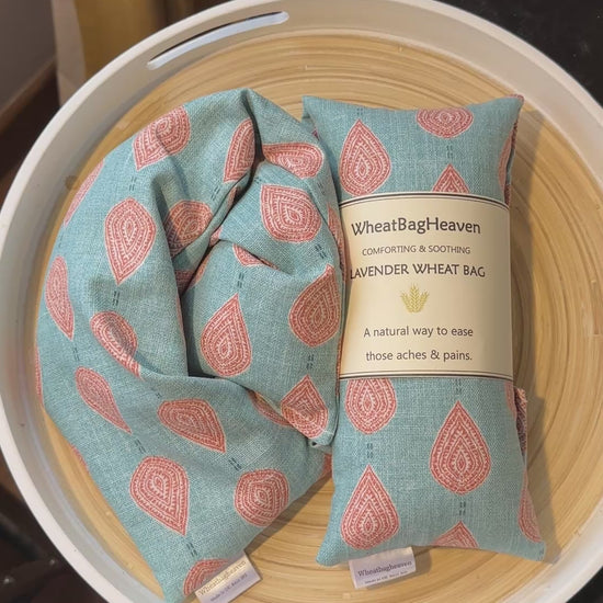 short video of A lavender wheat bag on a wooden tray. The wheat bag has a paisley teardrop pattern in shades of green and cream with a base colour of ochre. Its labelled "WheatBagHeaven" comforting and soothing lavender wheat bag. A natural way to ease those aches and pains. Also a tag that reads WheatBagHeaven