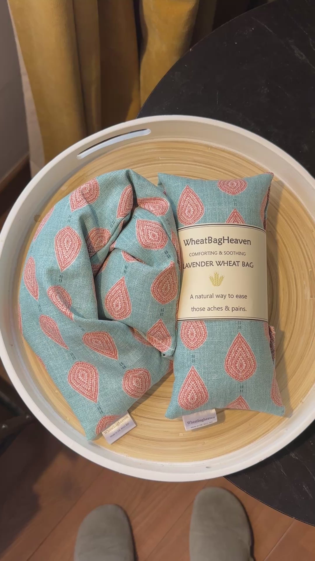 short video of A lavender wheat bag on a wooden tray. The wheat bag has a paisley teardrop pattern in shades of green and cream with a base colour of ochre. Its labelled "WheatBagHeaven" comforting and soothing lavender wheat bag. A natural way to ease those aches and pains. Also a tag that reads WheatBagHeaven